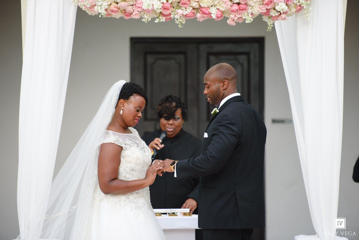 Bride and groom exchange rings during outdoor wedding ceremony beneath floral arch with officiant.