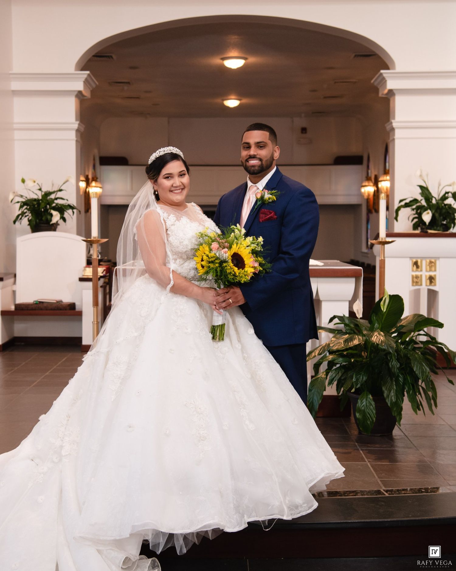 Bride in white ballgown with sunflower bouquet and groom in navy suit pose together inside a church.