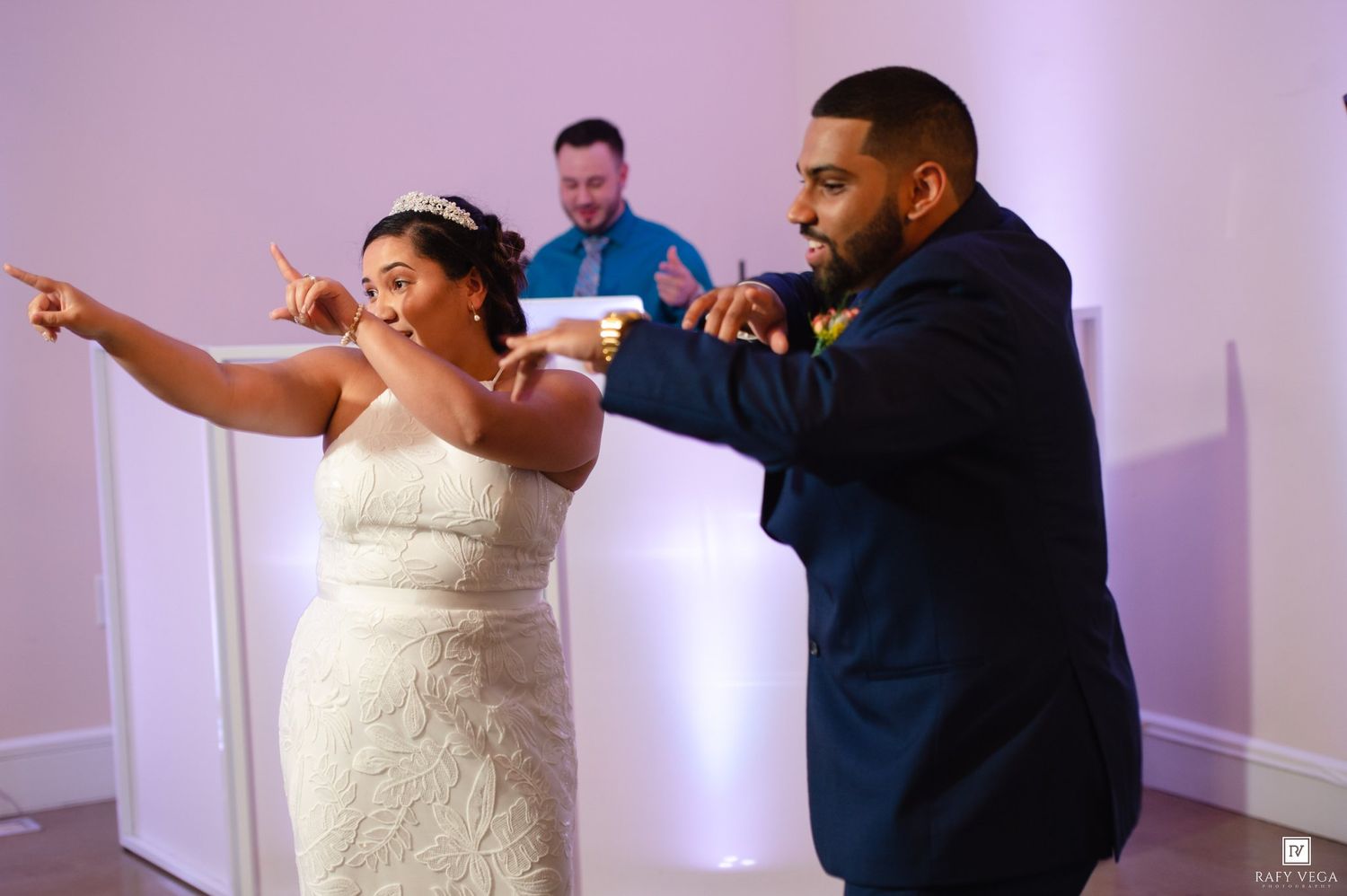 Bride in white gown and groom in navy suit dance joyfully at wedding reception with DJ in background.