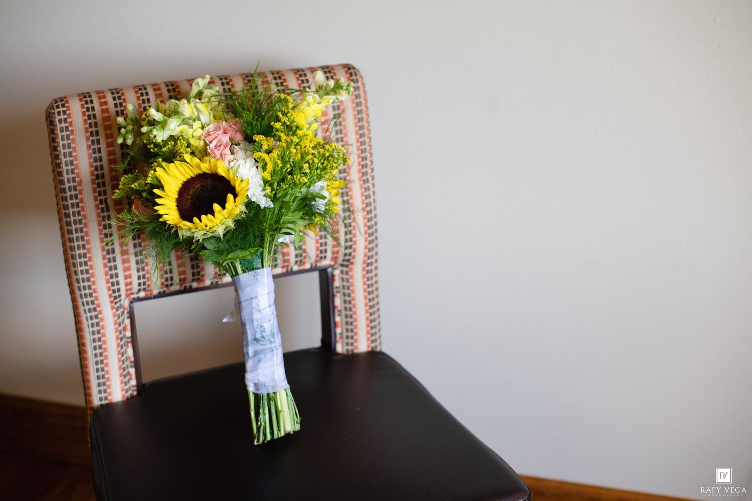 A vibrant wedding bouquet featuring a sunflower and mixed blooms resting on a vintage patterned chair.