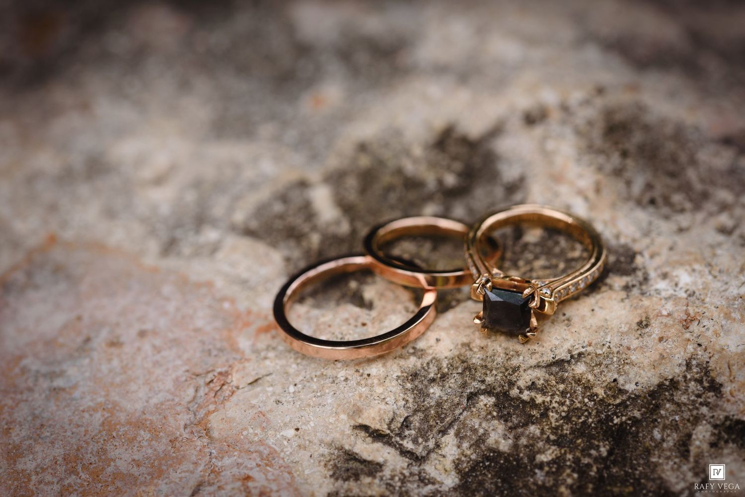 Three gold wedding rings with a sapphire engagement ring resting on a textured stone surface.