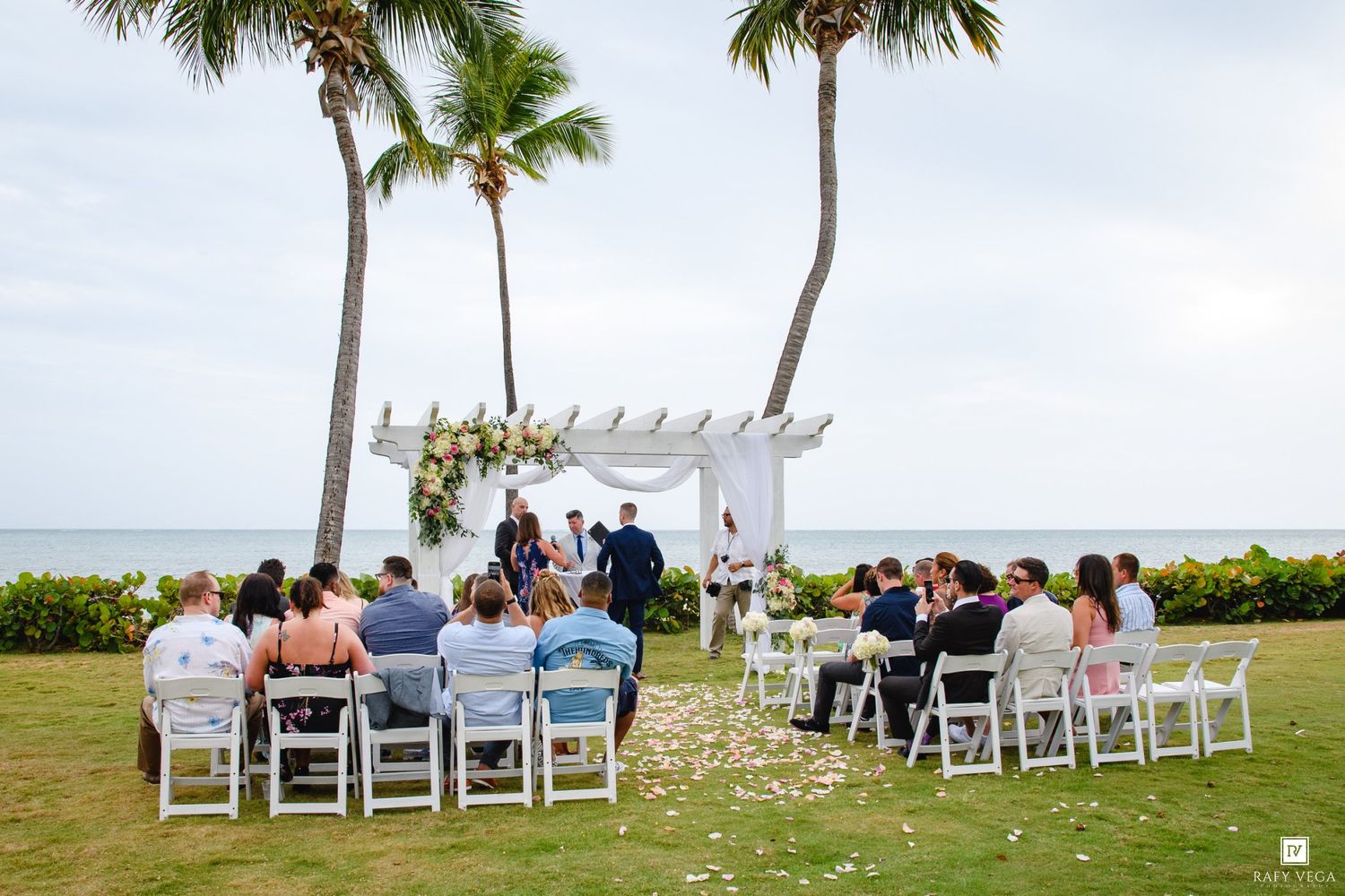 Intimate beachside wedding ceremony with guests seated facing floral arch between palm trees.