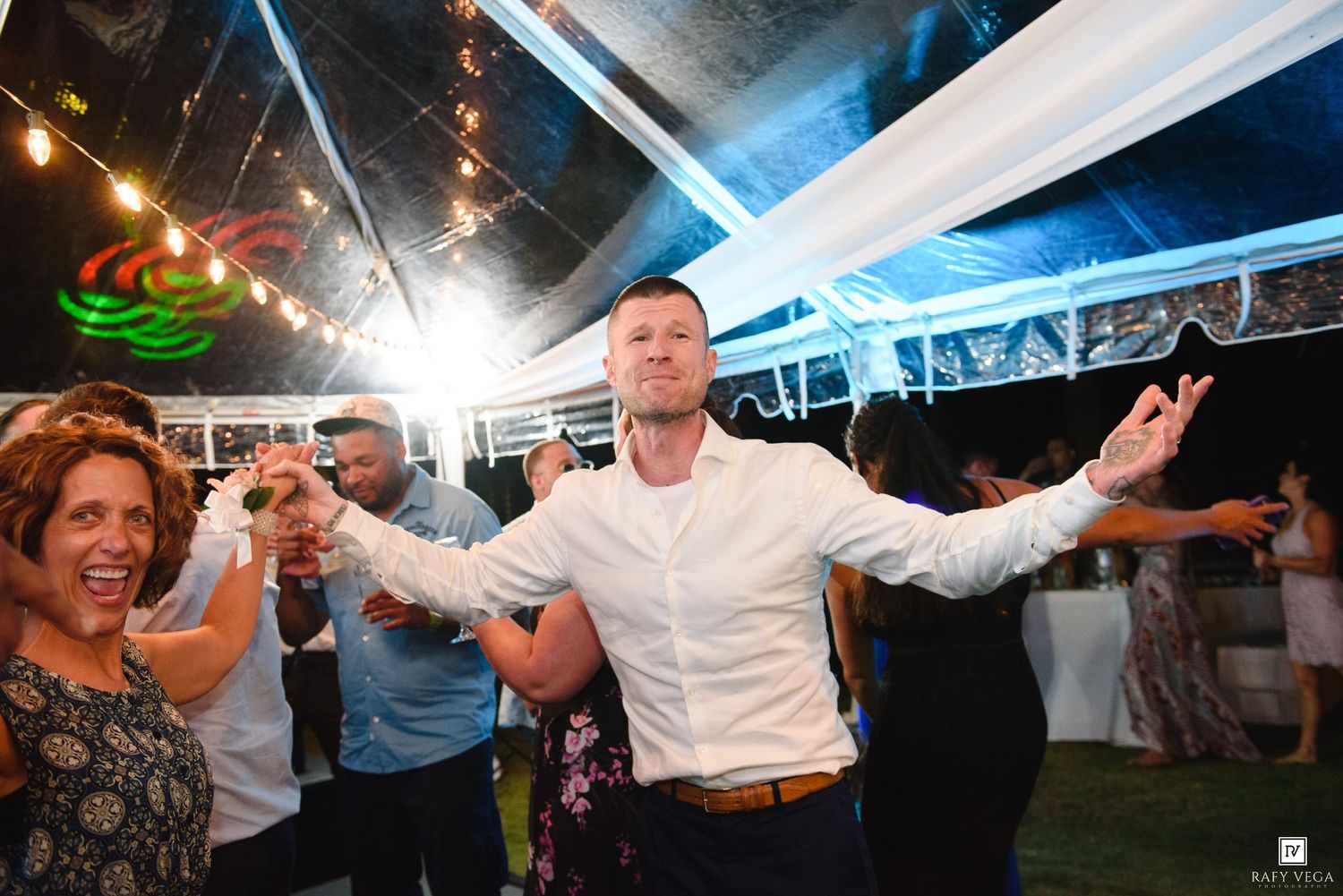 A man in a white shirt dances joyfully with arms outstretched at an outdoor wedding reception under a tent.