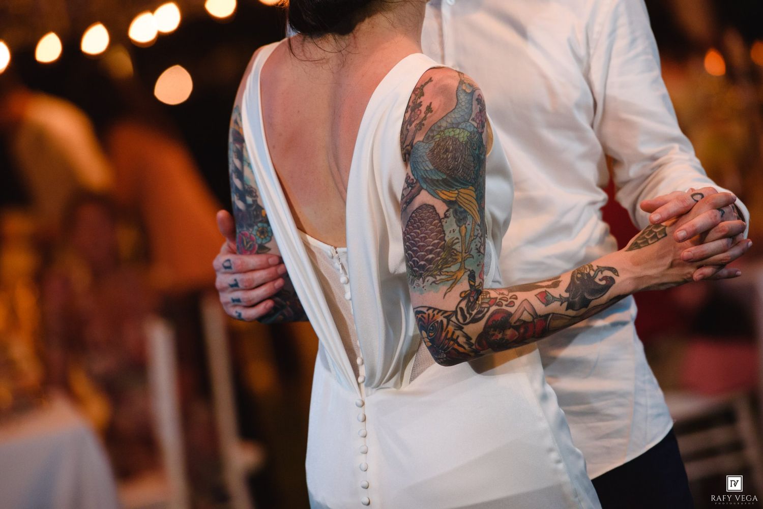 Tattooed couple sharing first dance at wedding reception, bride in backless white gown, groom with full sleeve tattoos.