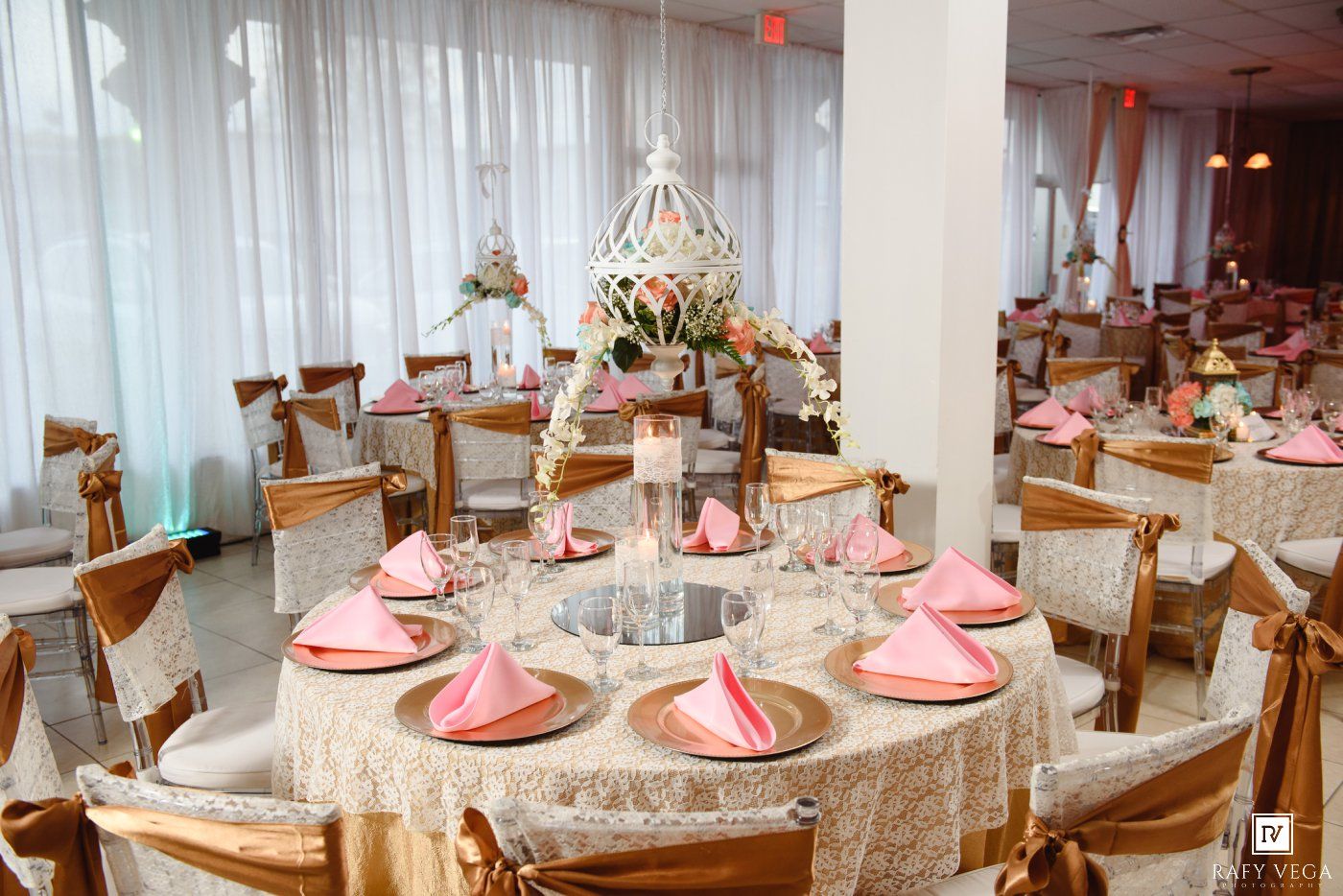 Elegant wedding reception hall with round tables, lace tablecloths, pink napkins, gold chairs, and floral centerpieces.