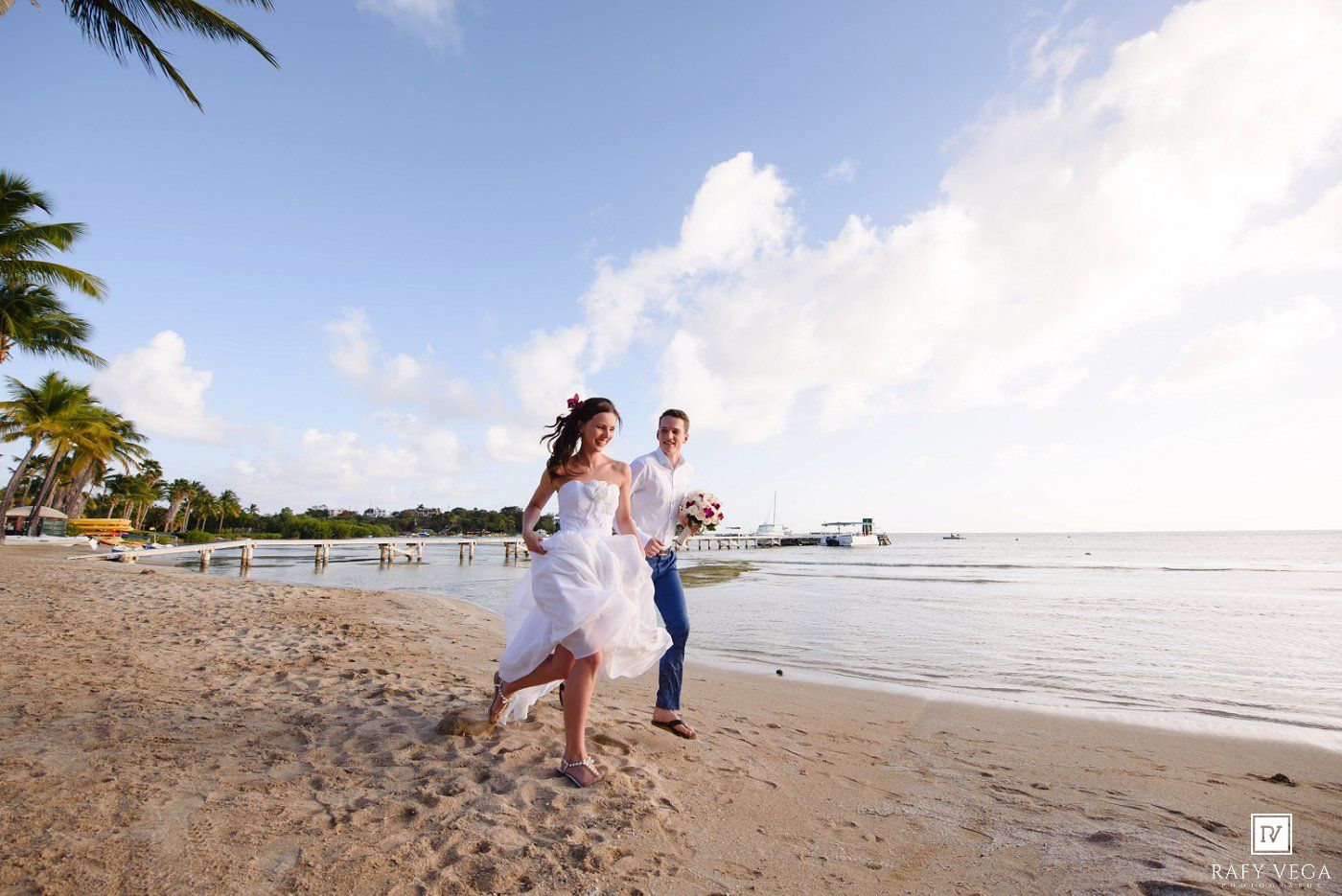 Bride and groom running joyfully on a tropical beach with palm trees and clear blue skies on their wedding day.