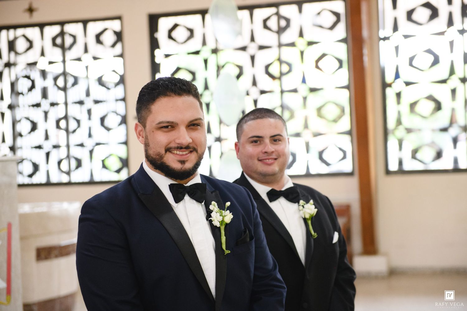 Two grooms in navy tuxedos with boutonnieres smile at a church wedding ceremony.