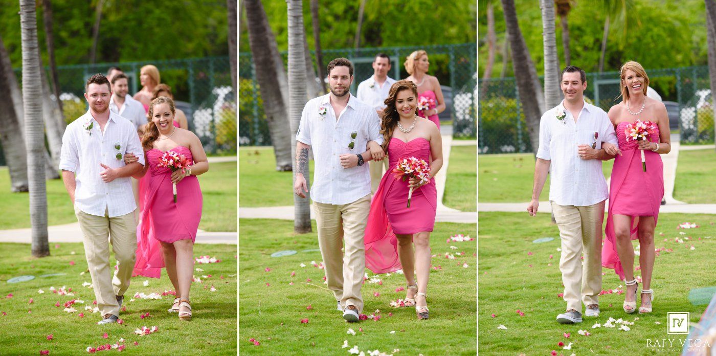 Bride and groom walk down aisle with wedding party in pink dresses on tropical lawn with palm trees.