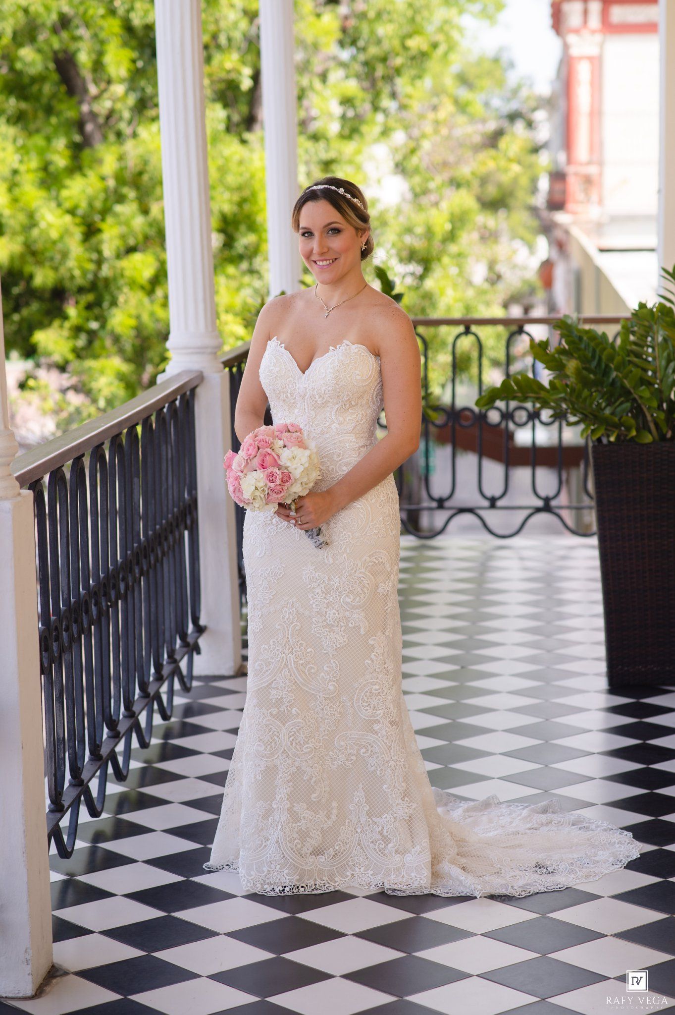 Bride in lace mermaid wedding gown holding pink bouquet on black and white checkered porch with white columns.