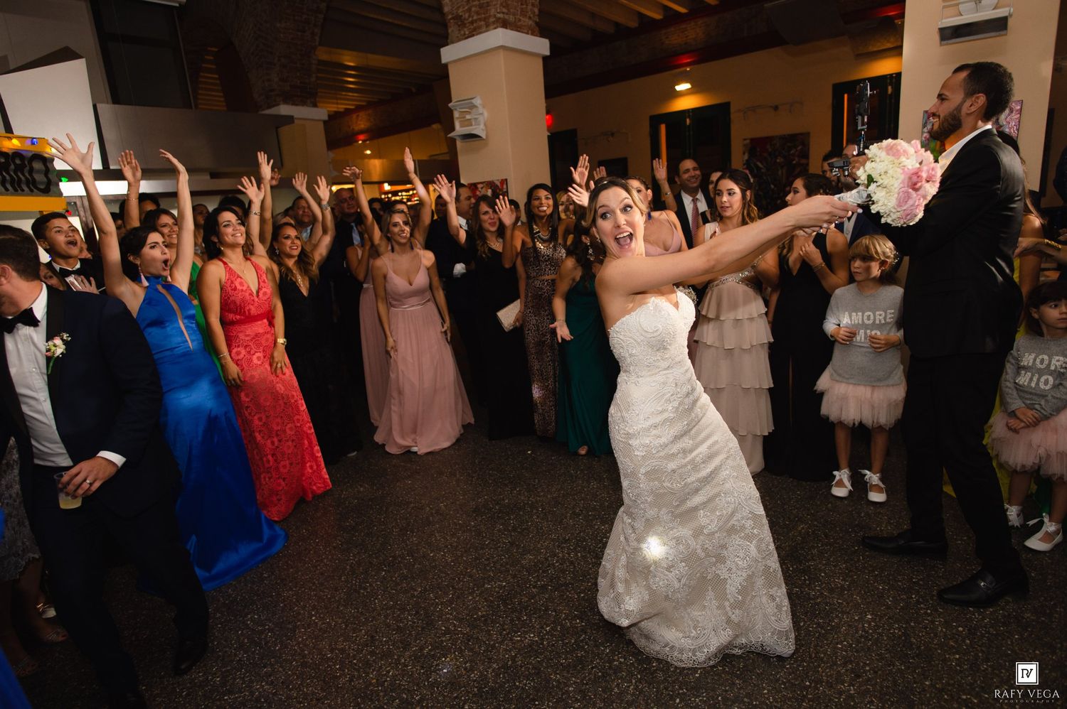 Bride in white gown tosses bouquet to excited guests at elegant indoor wedding reception celebration.