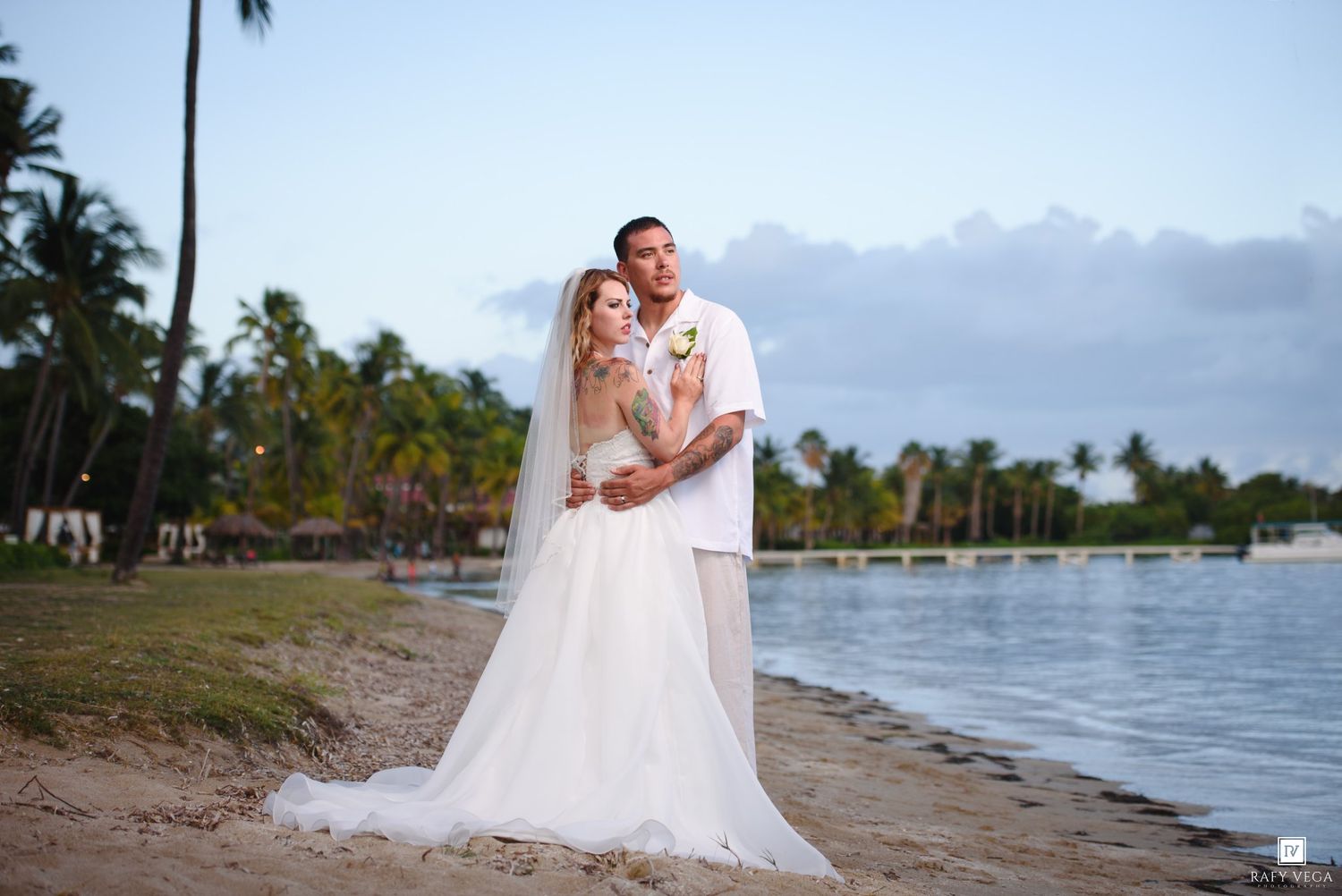 Bride and groom embrace on tropical beach at sunset, palm trees and calm water in background.