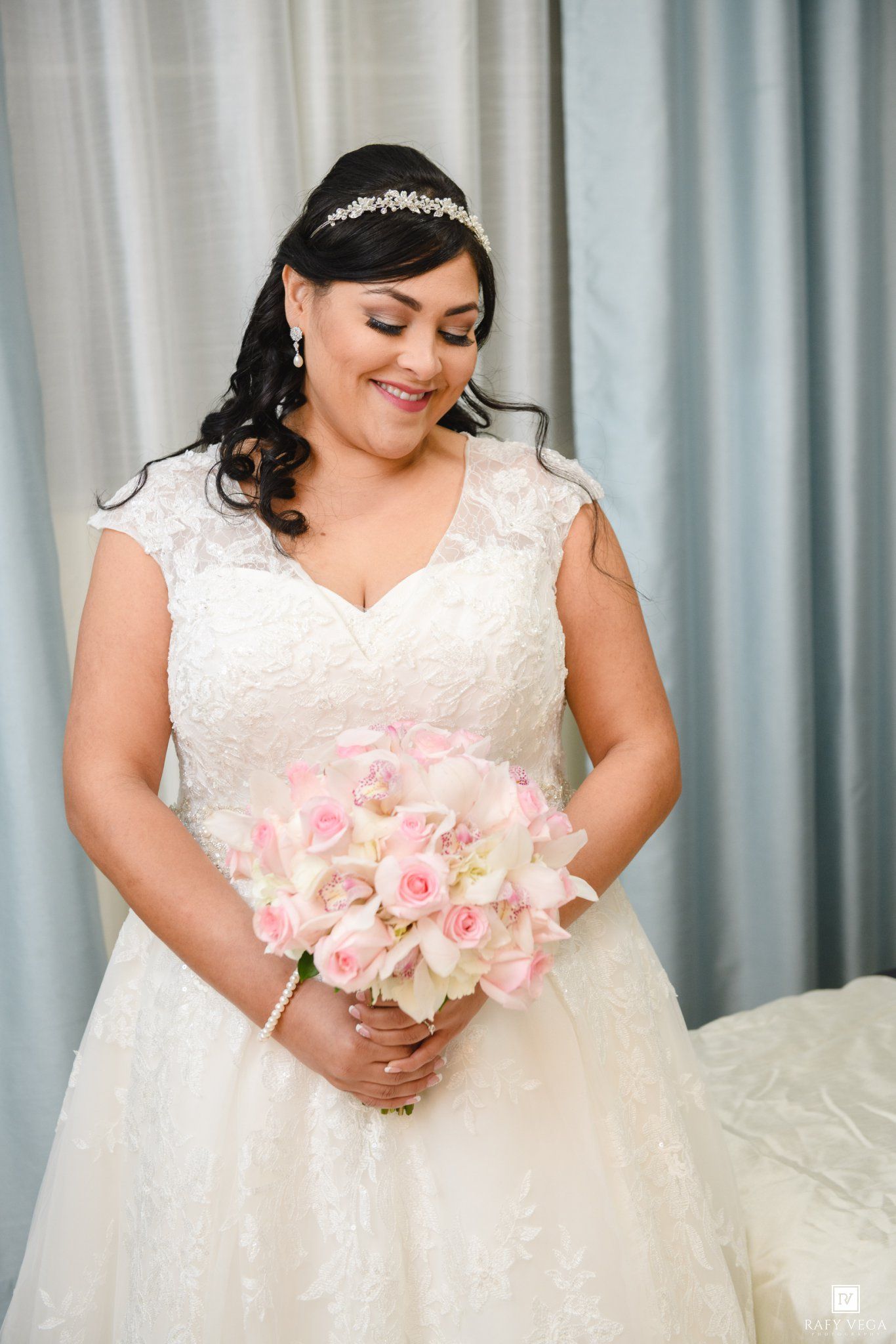 Smiling bride in white lace gown holding pink rose bouquet, wearing pearl jewelry and crystal tiara.