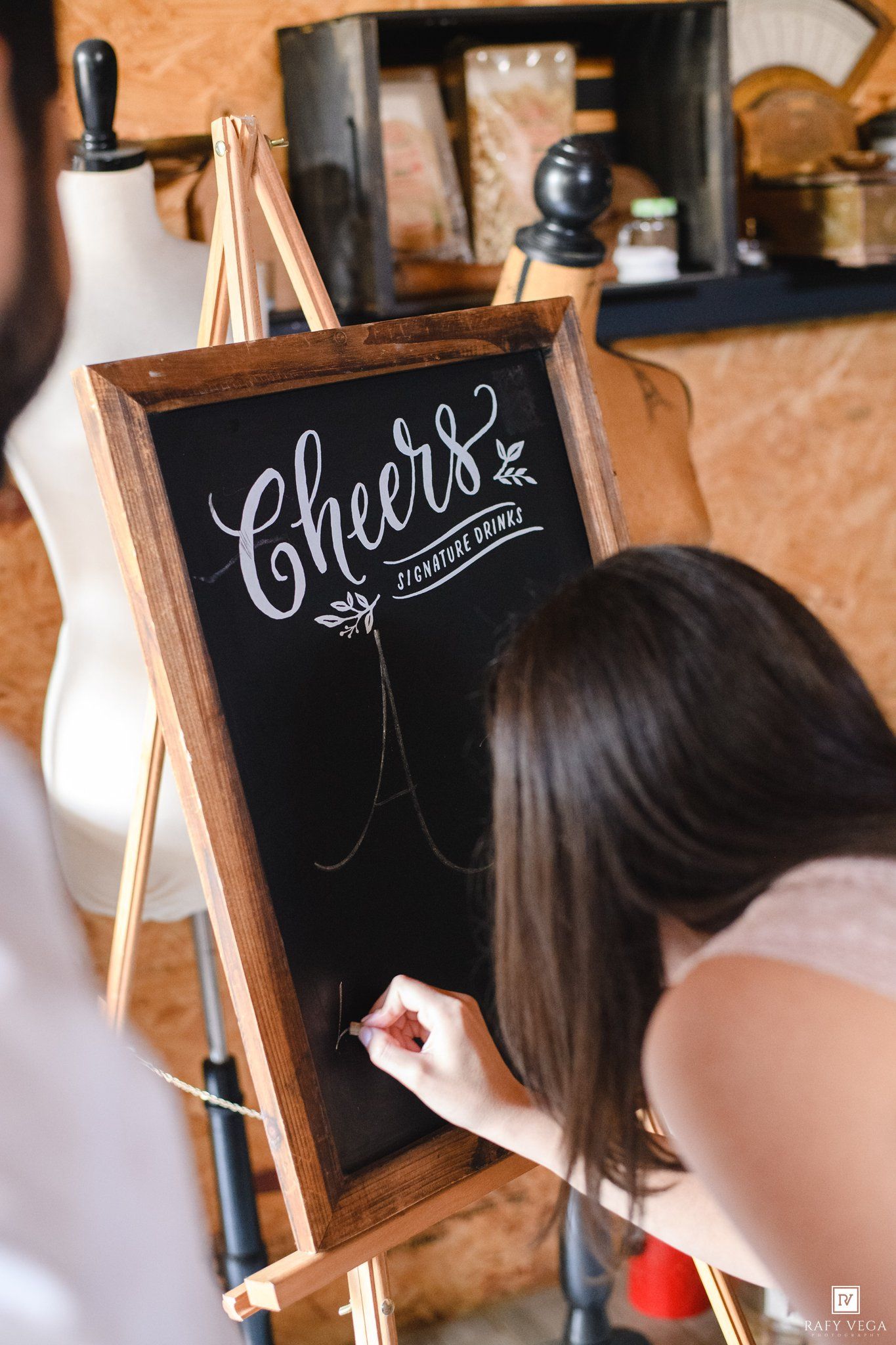 Woman writing in chalk calligraphy on a black chalkboard sign reading Cheers Signature Drinks on a wooden easel.