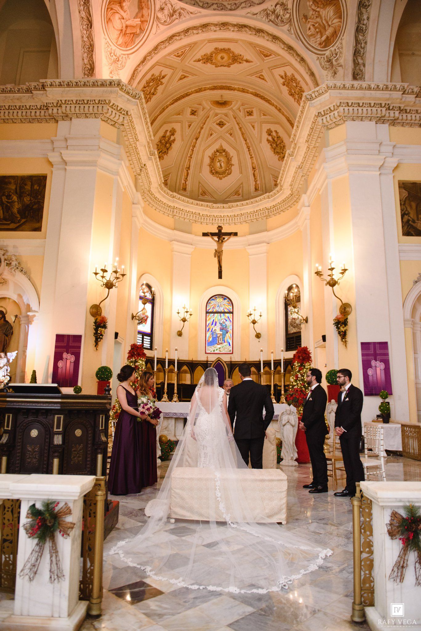 Elegant Catholic church wedding ceremony with bride in long veil, groom, and wedding party at ornate altar.