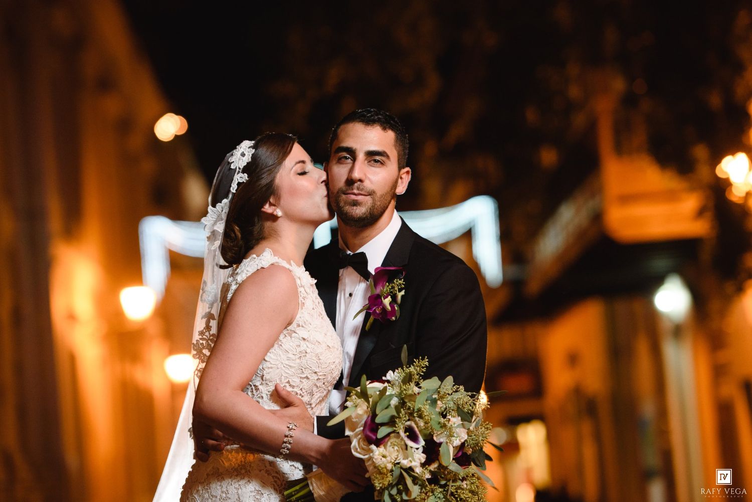 Bride and groom share romantic moment at night, she holds floral bouquet, city lights glowing warmly behind them.