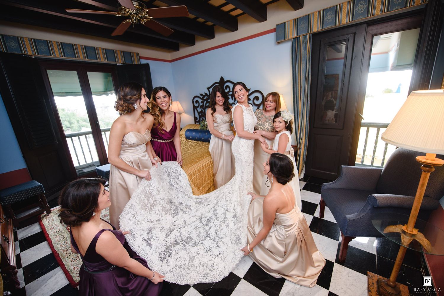 Bridesmaids helping bride with her lace wedding gown in an elegant room with checkered floors.