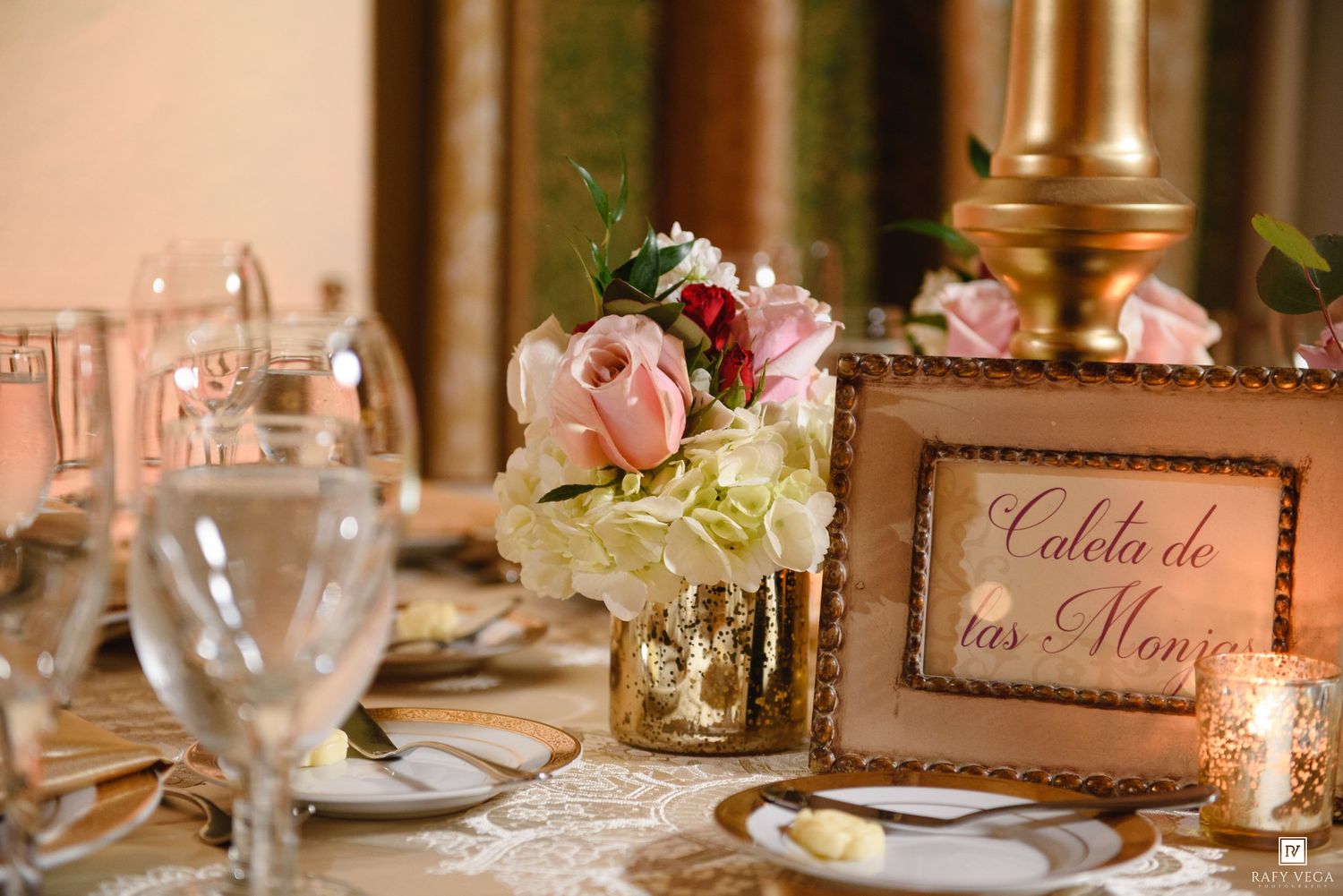Elegant wedding reception table with pink roses, hydrangeas in gold vase, crystal glasses, and decorative place card sign.