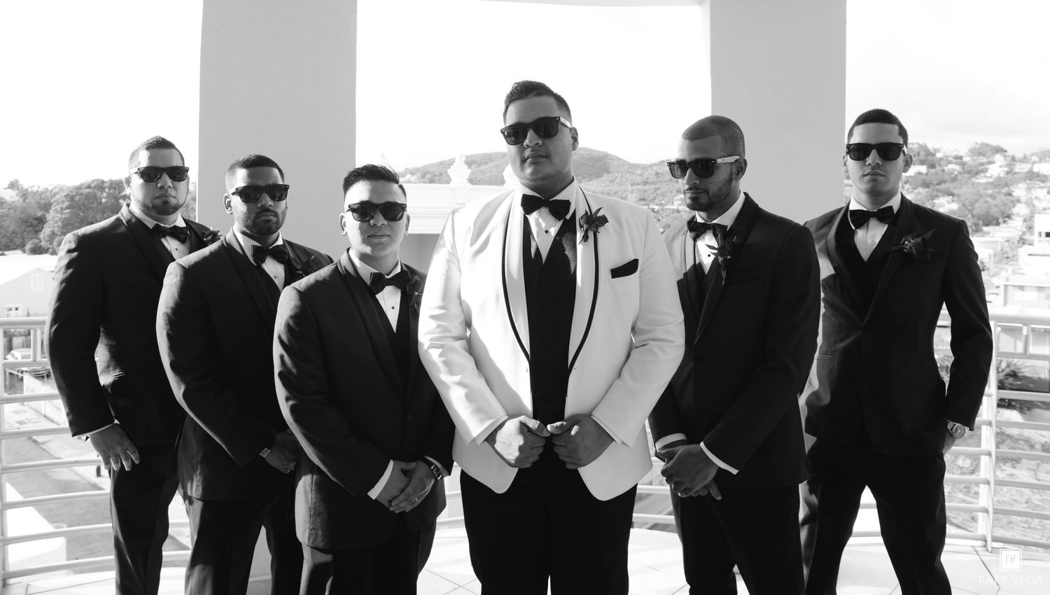Groom in white tuxedo poses with groomsmen in black suits and sunglasses on outdoor venue balcony.