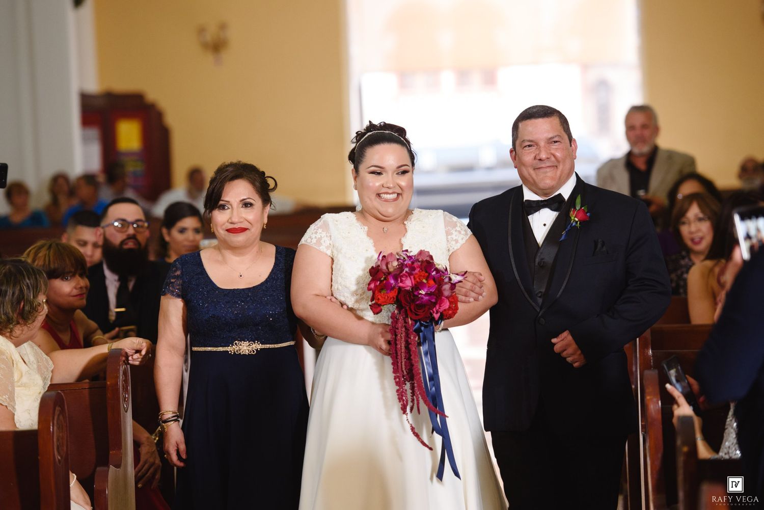 Bride in white gown walks down church aisle with parents during wedding ceremony, holding red and blue bouquet.