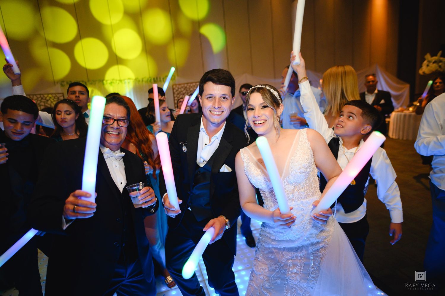 Bride and groom celebrate at wedding reception with guests holding glowing light sticks on the dance floor.