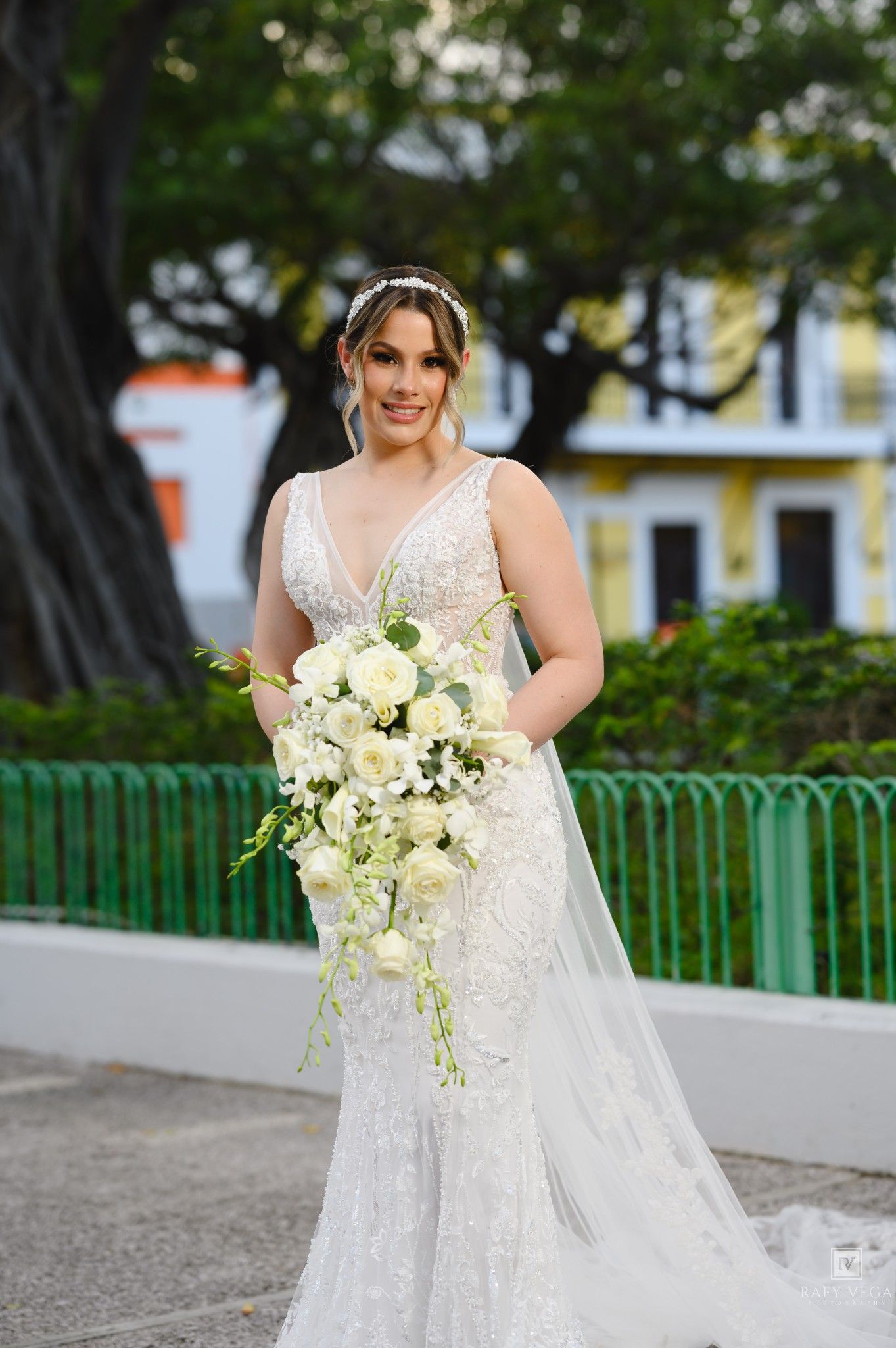 Bride in beaded mermaid gown holding white cascading bouquet outdoors near green fence and large tree.