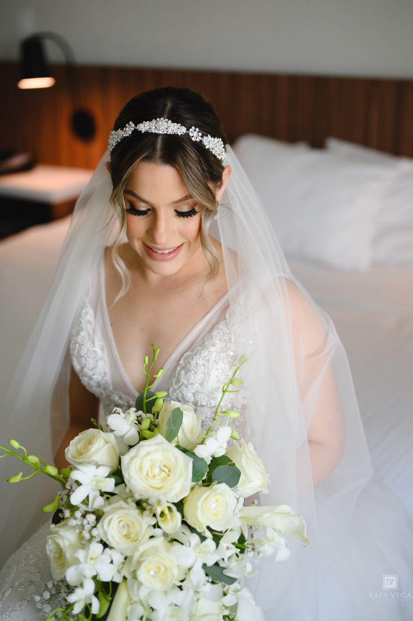 Bride in lace wedding gown and veil holds white rose bouquet, smiling down in elegant bridal portrait.