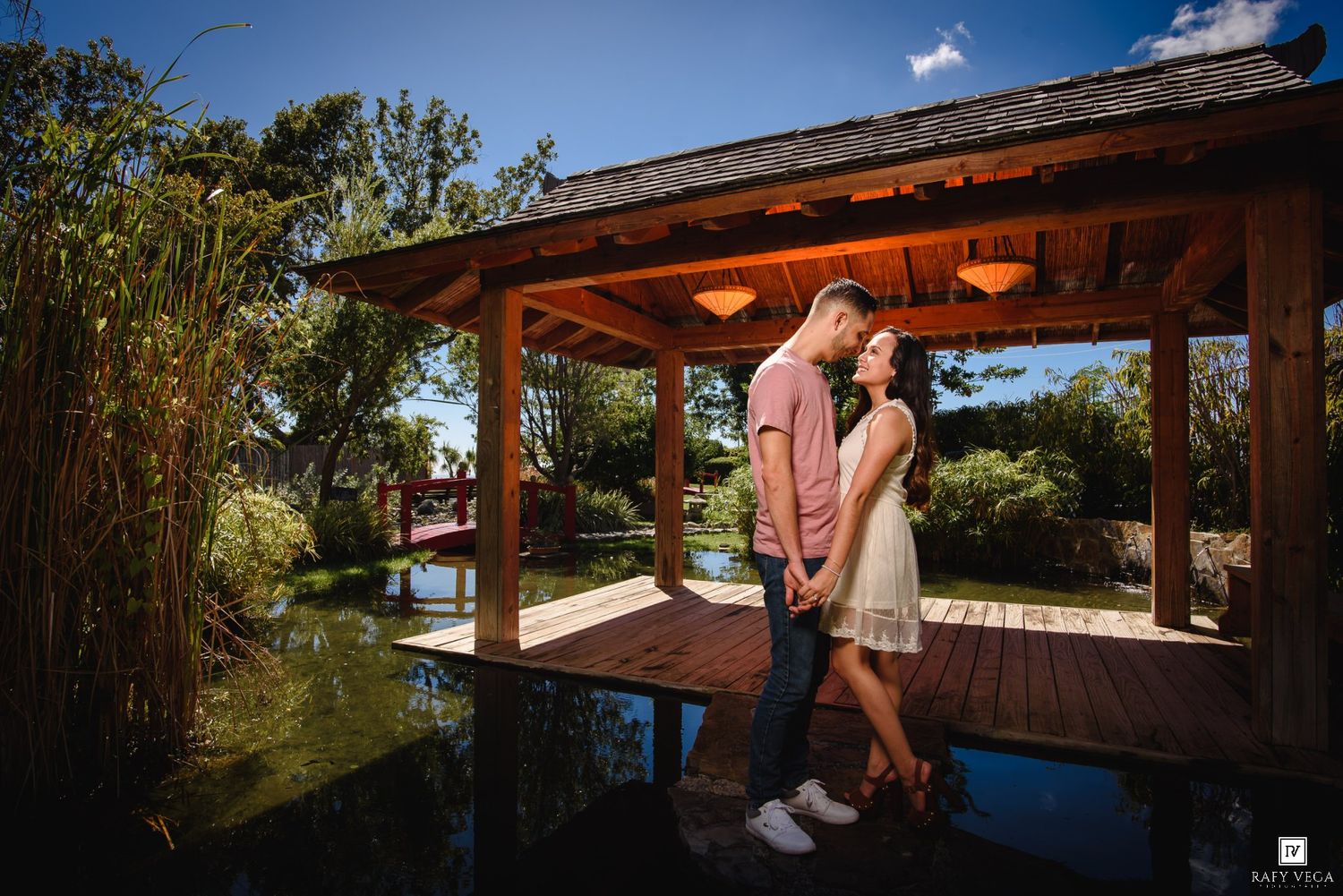 Couple holding hands under a wooden gazebo by a serene pond in a lush garden on a sunny day.
