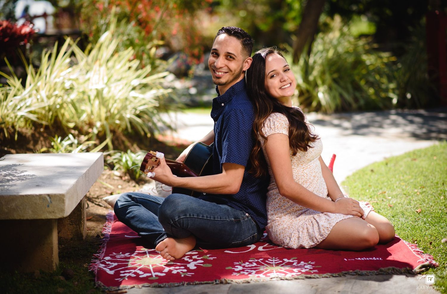 Happy couple sitting back-to-back on a red blanket in a sunny garden, smiling and relaxed outdoors.