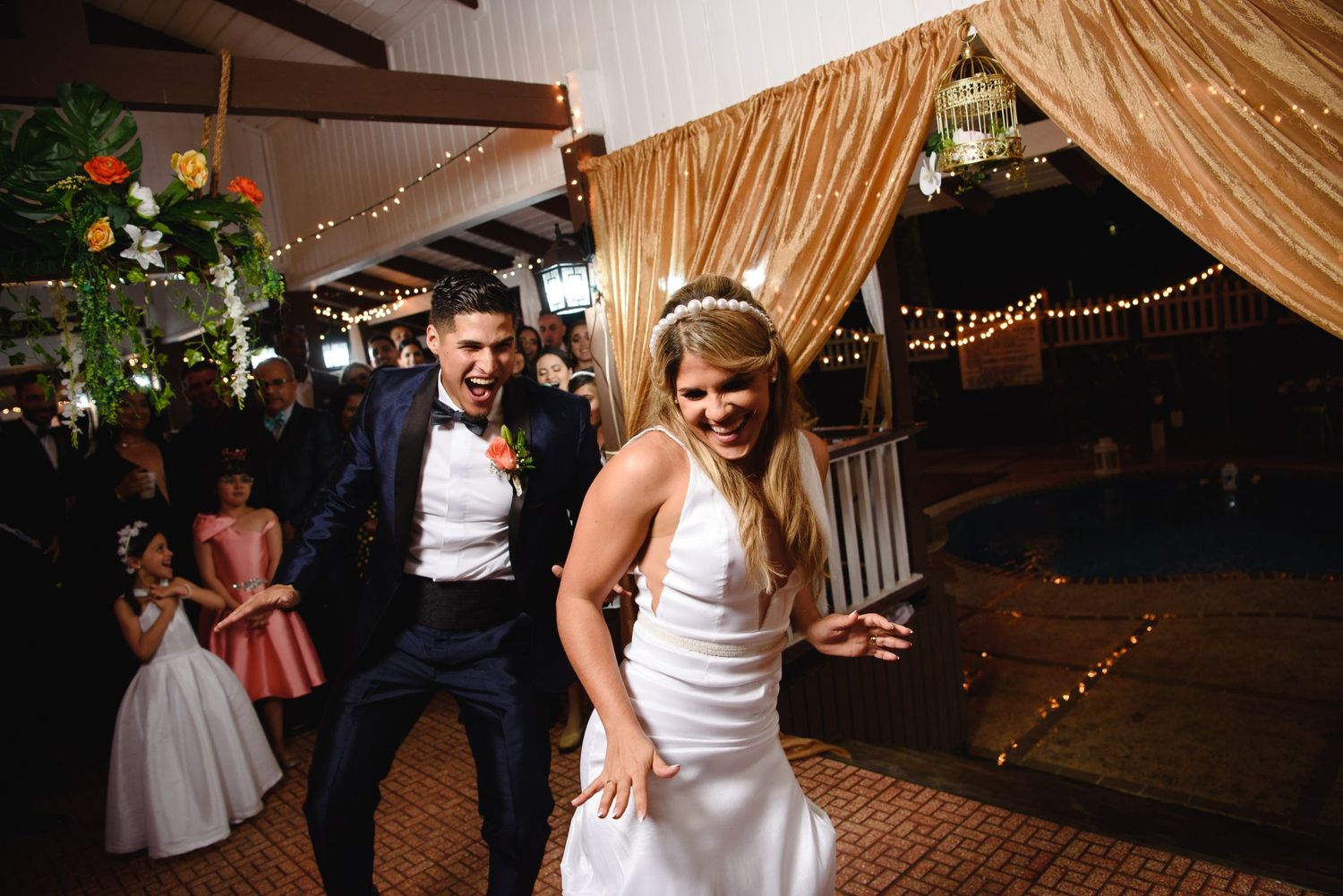 Happy bride and groom laughing during first dance at wedding reception with string lights and gold curtains.