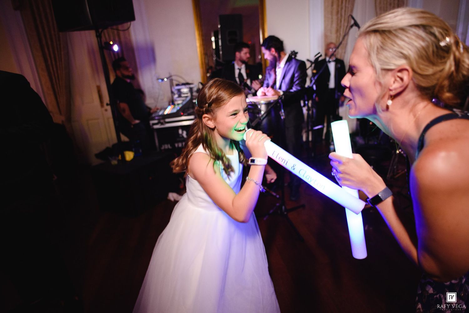 Young girl in white dress singing with woman at wedding reception with live band performing in background.