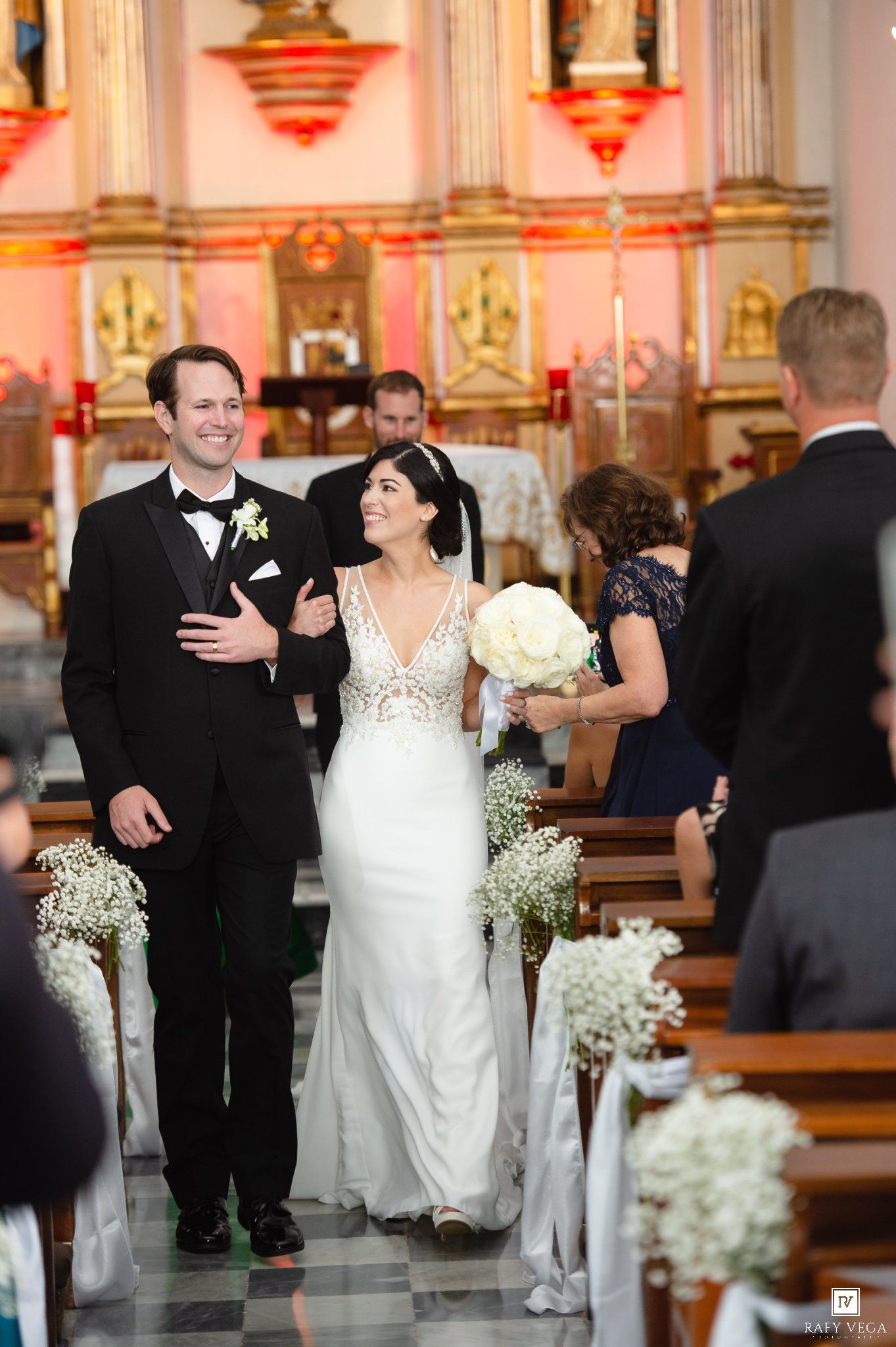 Bride and groom walk down church aisle after wedding ceremony, bride holding white bouquet in elegant white gown.
