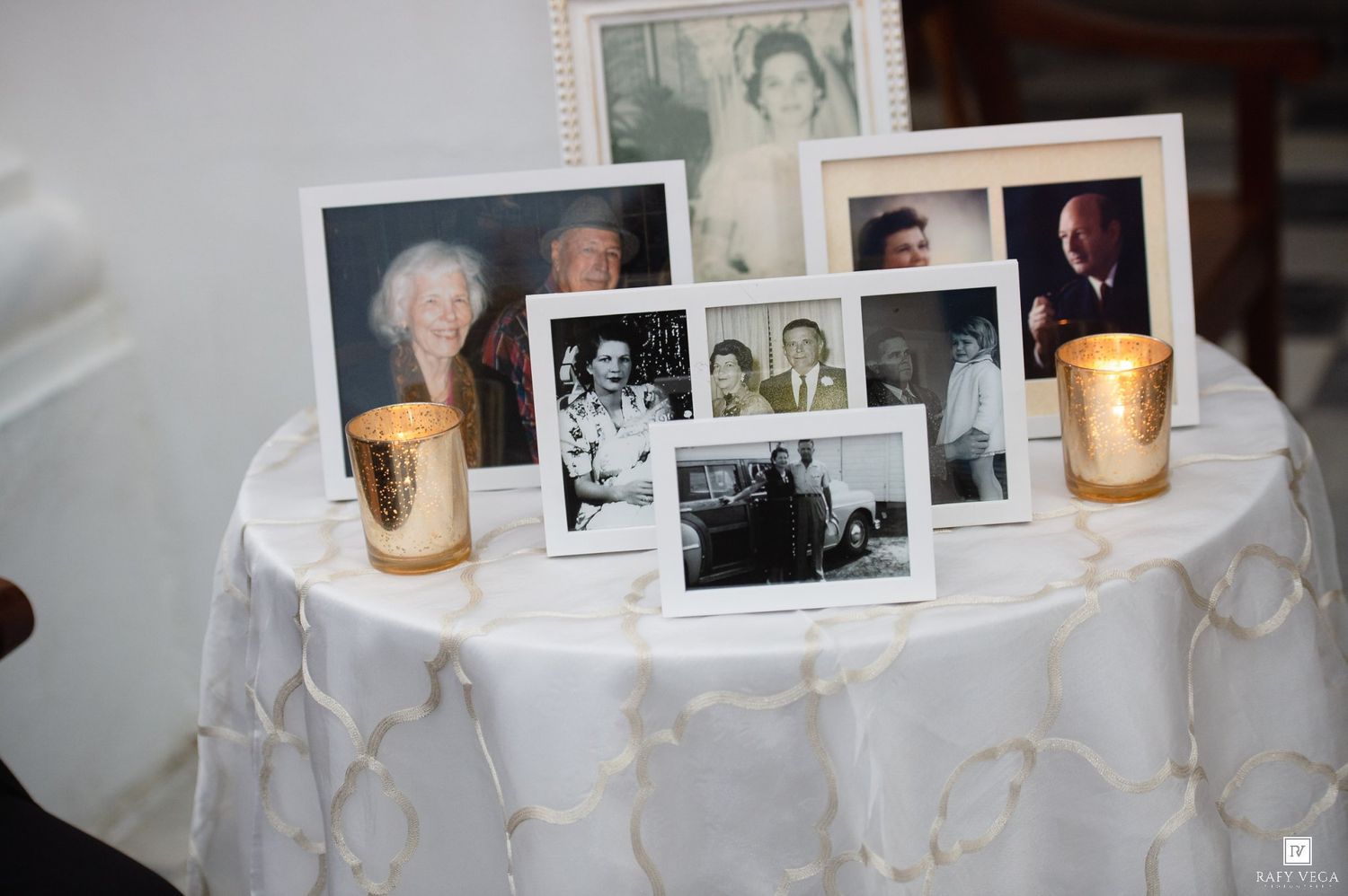 A memorial table with black-and-white family photos and gold candle holders on a white embroidered tablecloth.