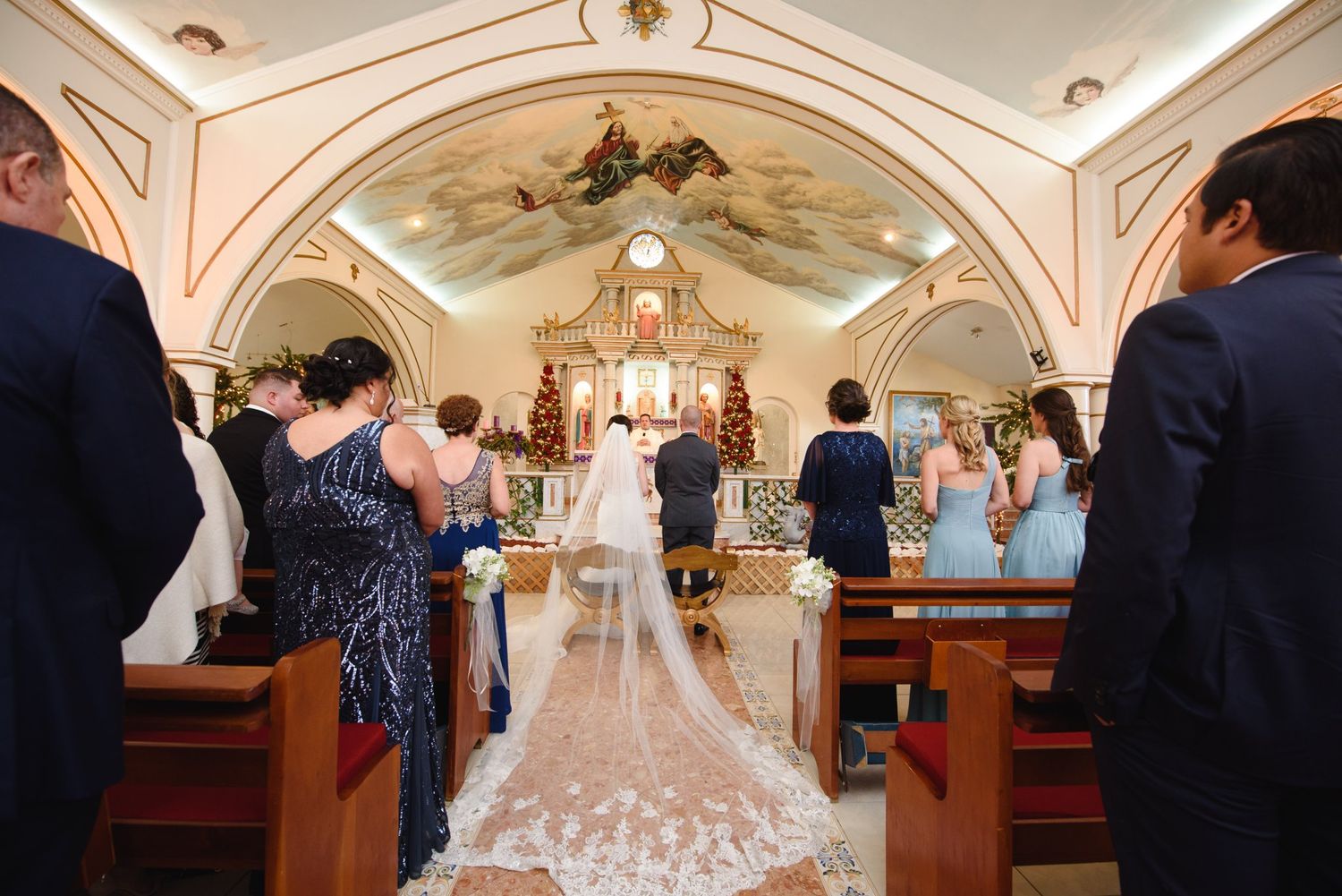 Bride walking down the aisle in a Catholic church with a long lace veil, surrounded by wedding party and guests.