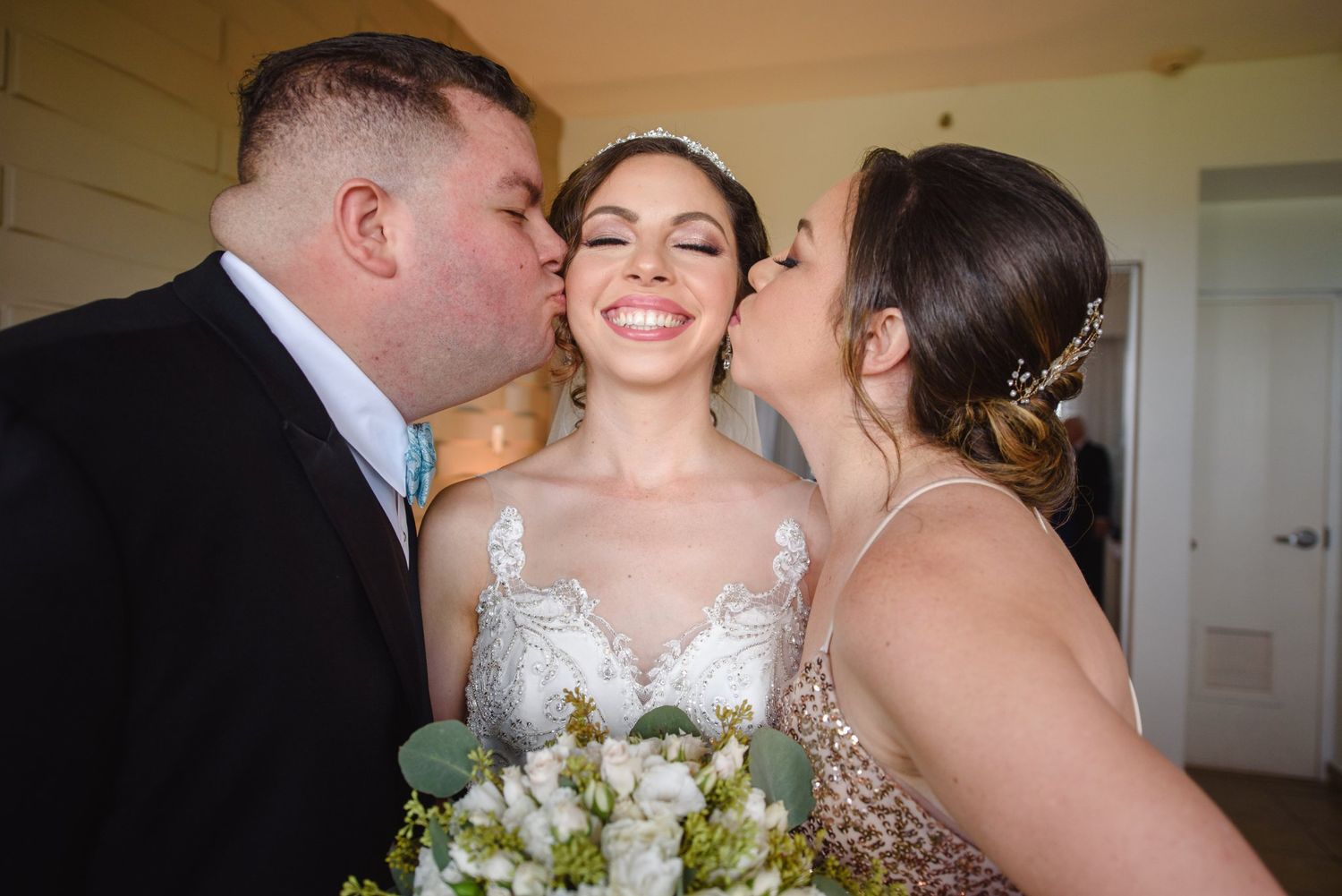 Bride smiling joyfully as groom and bridesmaid kiss her cheeks, holding white floral bouquet at wedding.