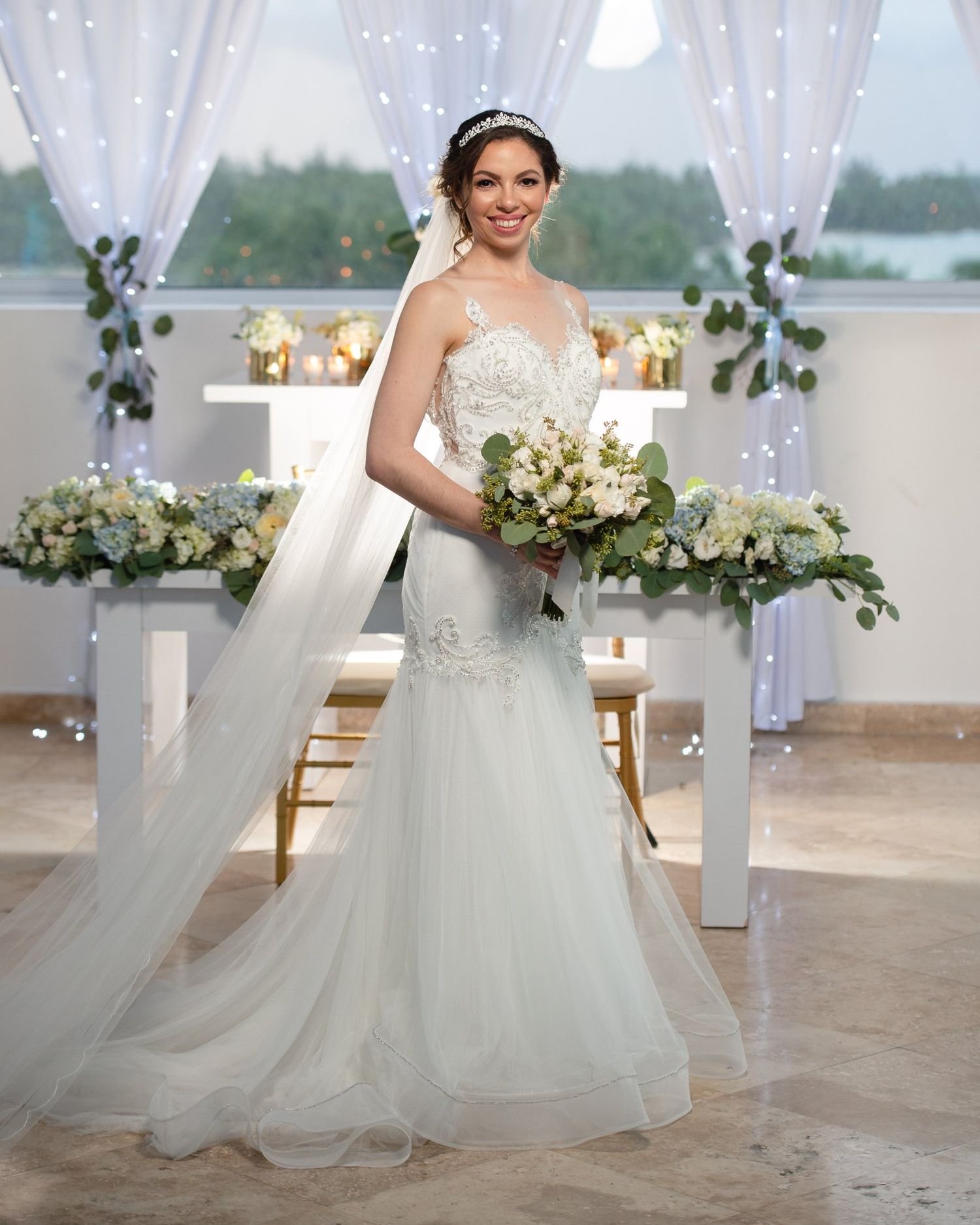 Bride in mermaid lace wedding dress holding bouquet at elegant ceremony altar with white drapes and floral arrangements.