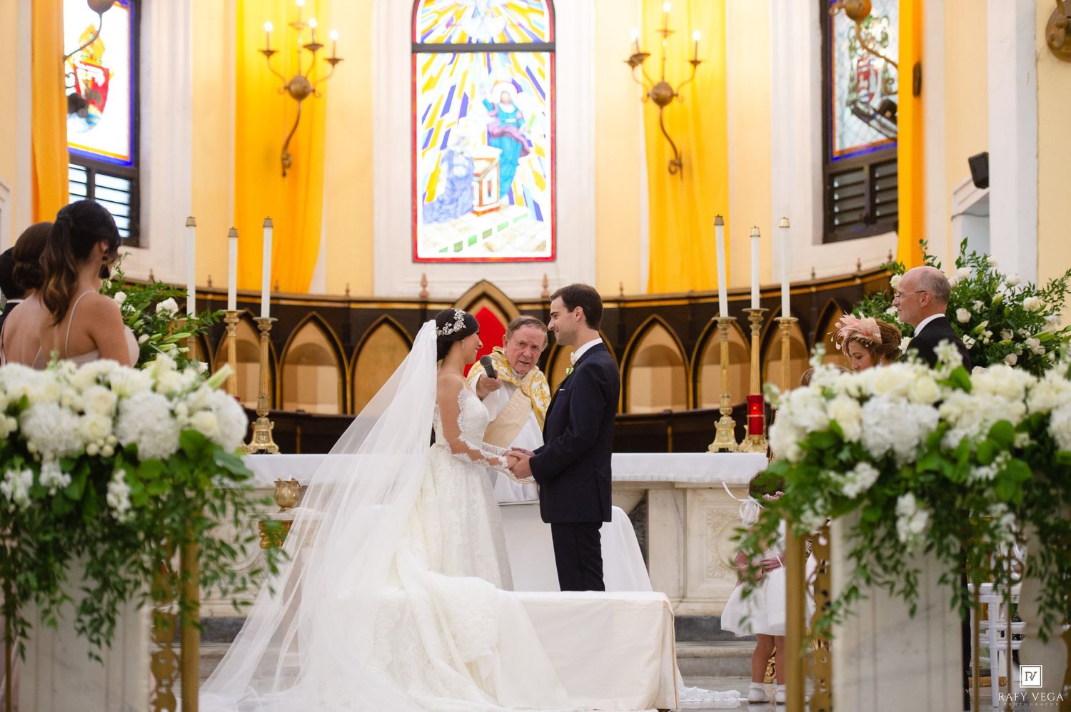 Bride and groom exchange vows at elegant church altar adorned with white floral arrangements and golden candlesticks.
