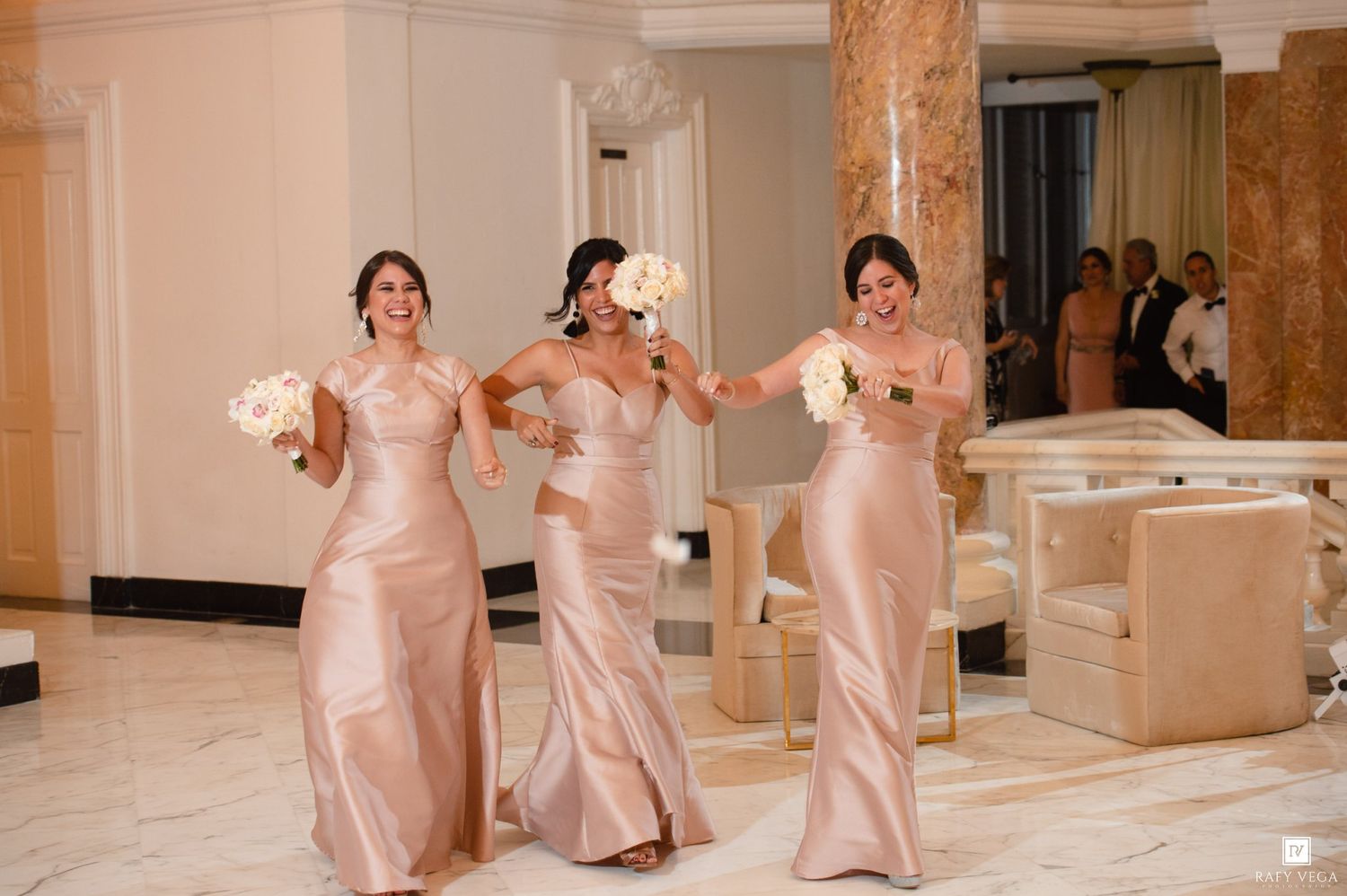 Three bridesmaids in rose gold satin gowns holding white bouquets walking through a luxurious marble venue.