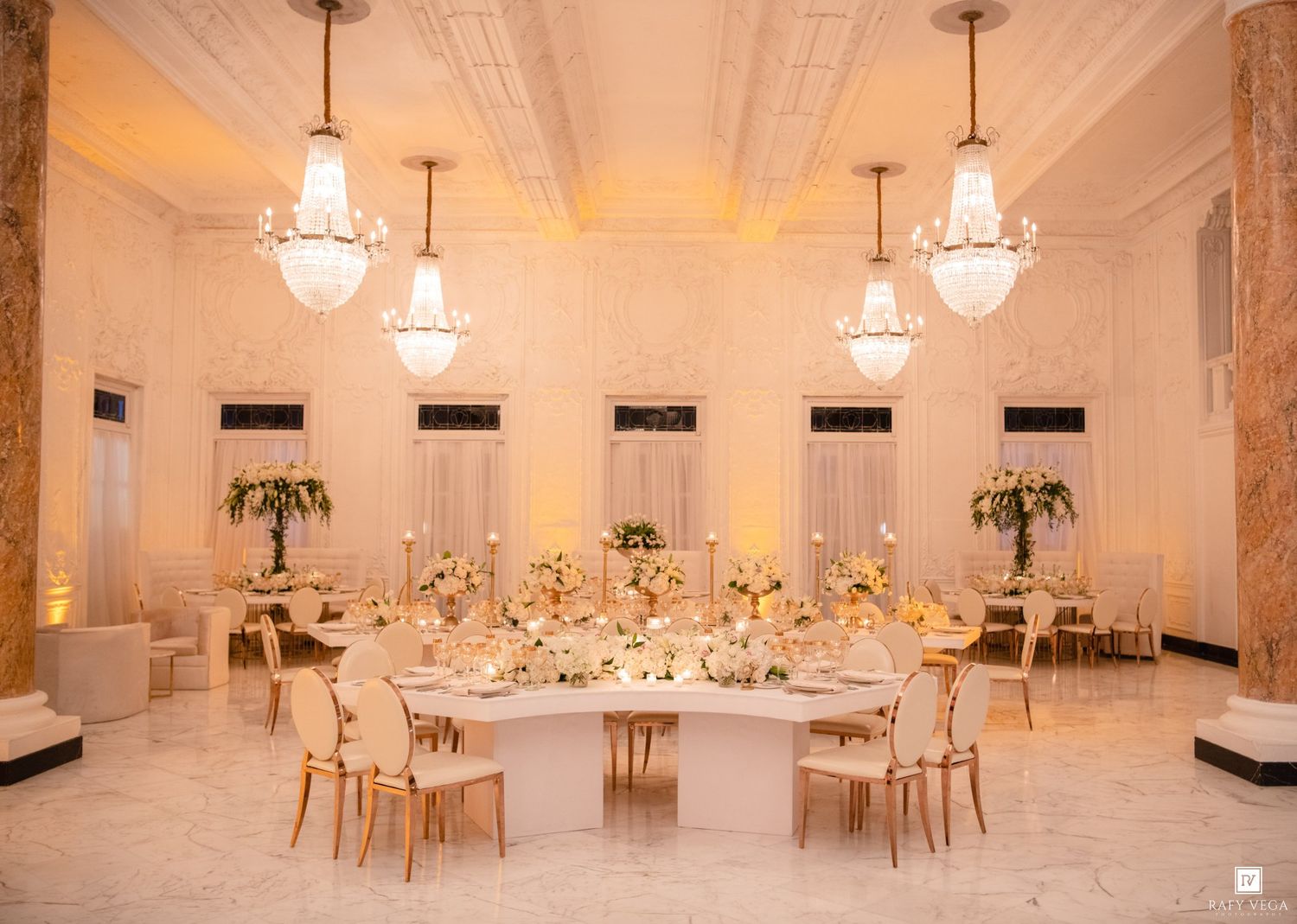 Elegant wedding reception ballroom with crystal chandeliers, gold chairs, and floral centerpieces on round tables with white linens.