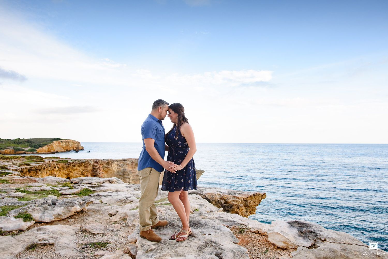 Couple sharing a romantic moment on rocky coastal cliffs with turquoise ocean and blue sky backdrop.