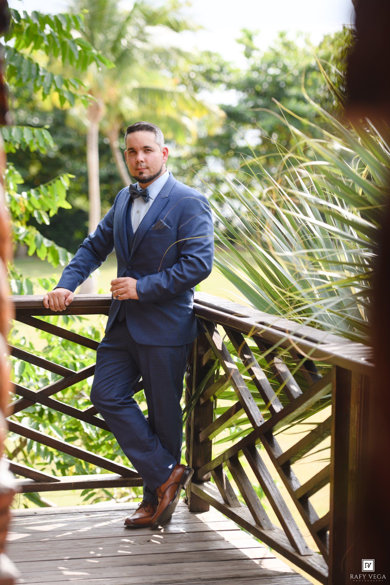 Man in navy blue suit with bow tie posing on wooden balcony surrounded by tropical greenery.