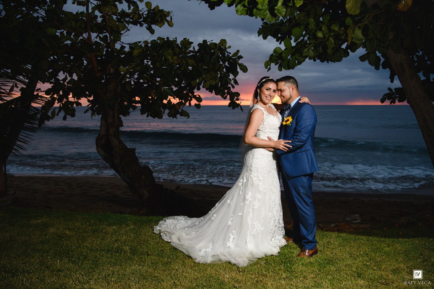 Bride and groom embrace at sunset by the ocean, with colorful pink and purple sky, lush greenery, and crashing waves.
