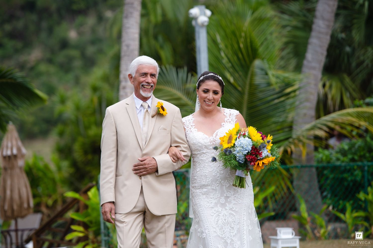 Bride in lace wedding dress walks with older man in beige suit, holding colorful bouquet, tropical palm trees background.