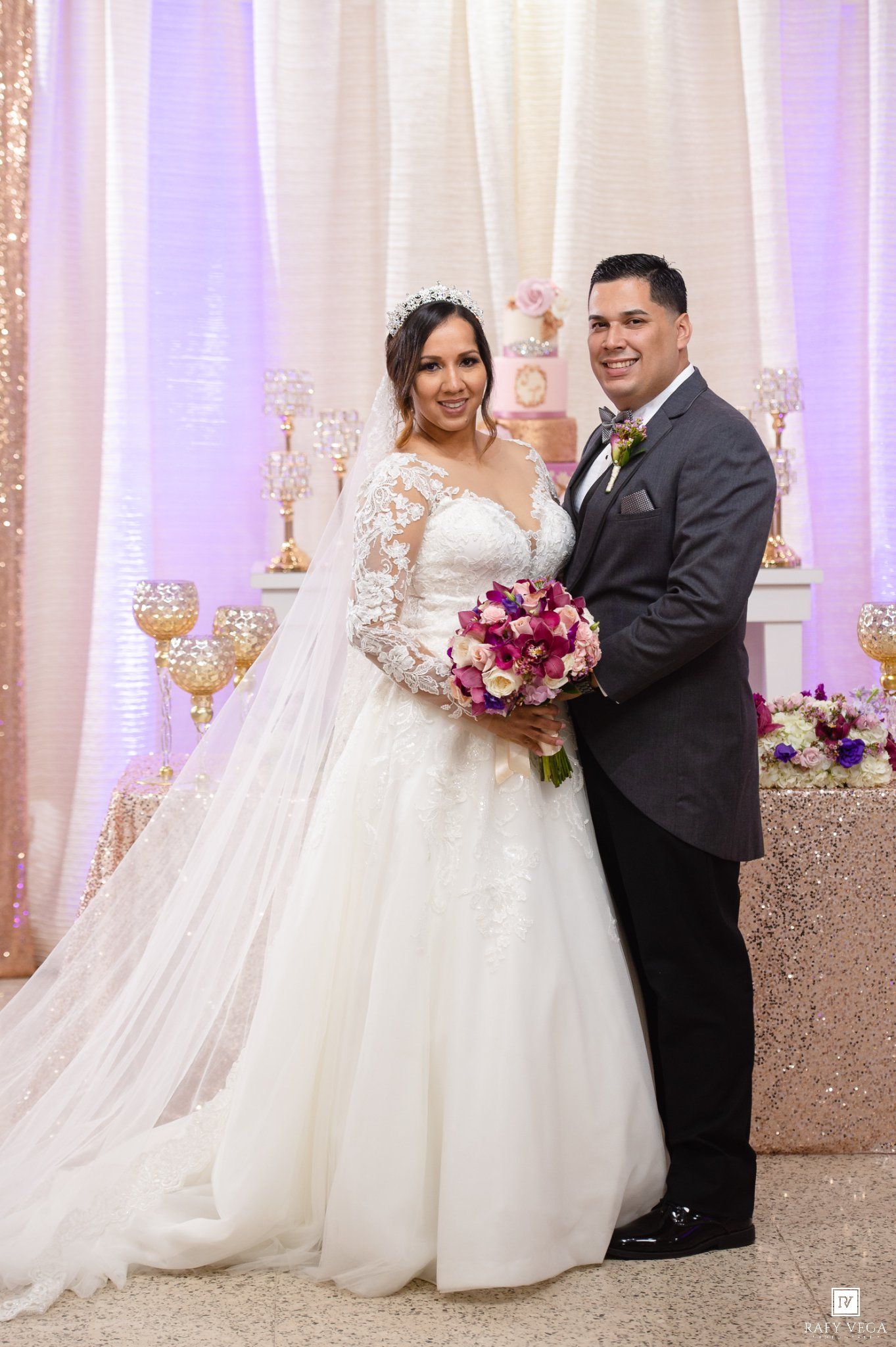 Bride in white lace gown holding purple bouquet stands with groom in gray suit before elegant wedding reception backdrop.
