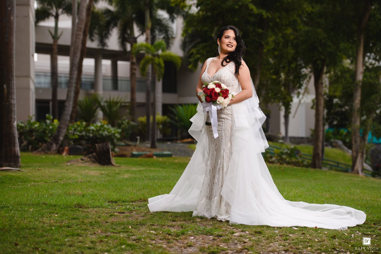 Bride in white wedding gown holding red bouquet poses outdoors on green lawn with tropical trees and building backdrop.