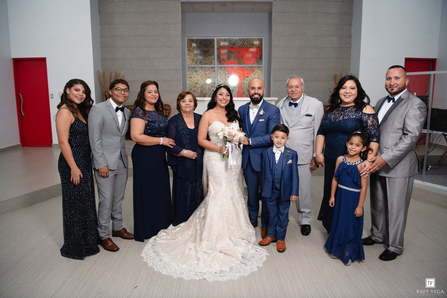 Wedding party poses together in navy blue formal attire, bride in white lace gown holding bouquet, groom in suit.