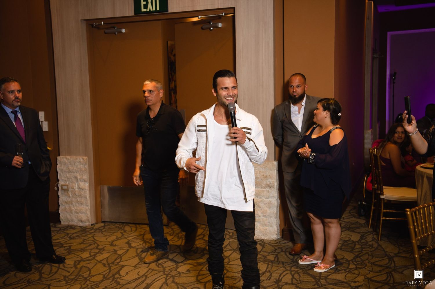 Man in white jacket speaks into microphone at elegant indoor event with guests and EXIT sign visible.