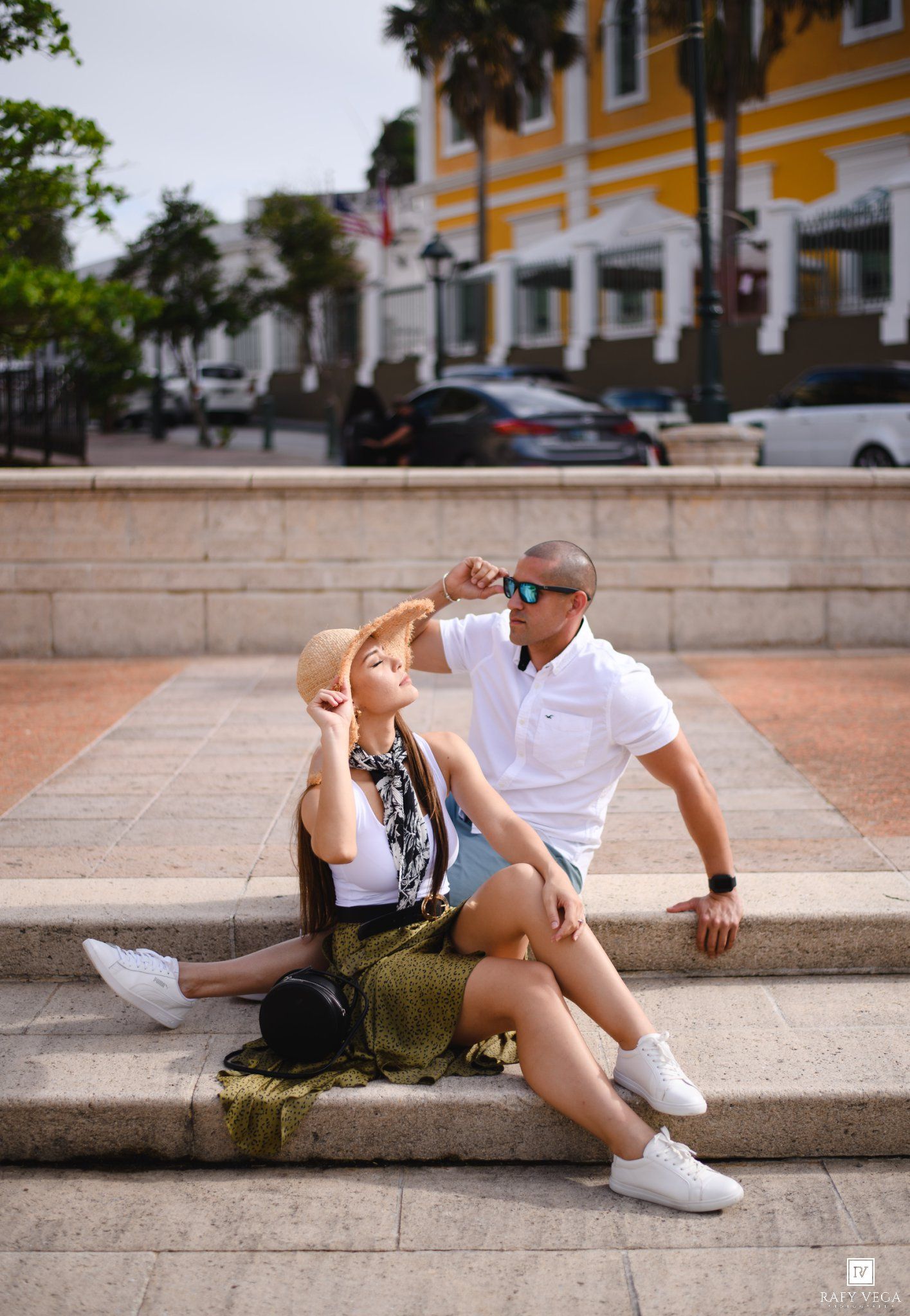 A couple sits on stone steps in a sunny urban plaza, woman in hat and man in white polo with sunglasses.