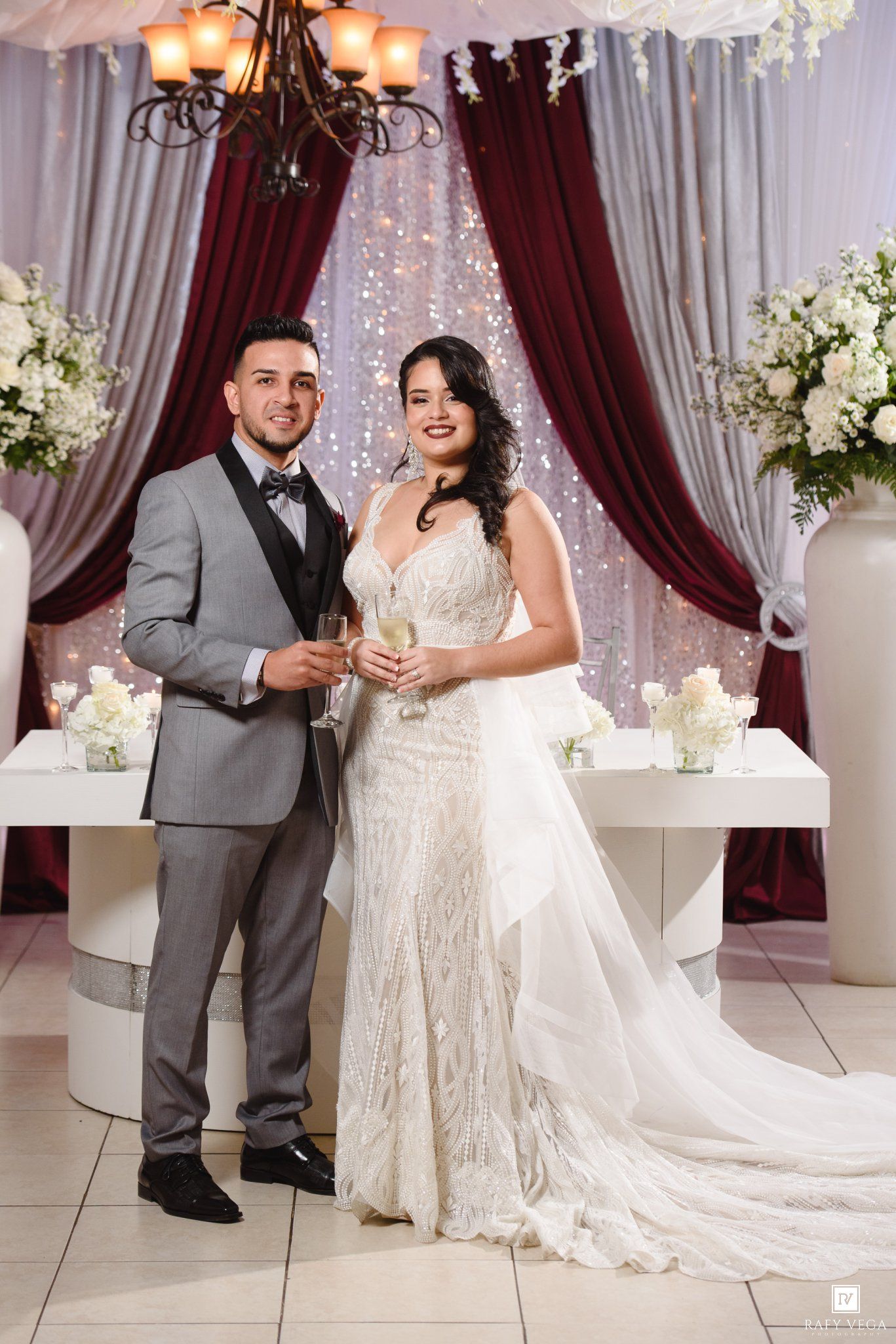 Bride and groom posing with champagne glasses at elegant wedding reception with burgundy drapes and white floral arrangements.