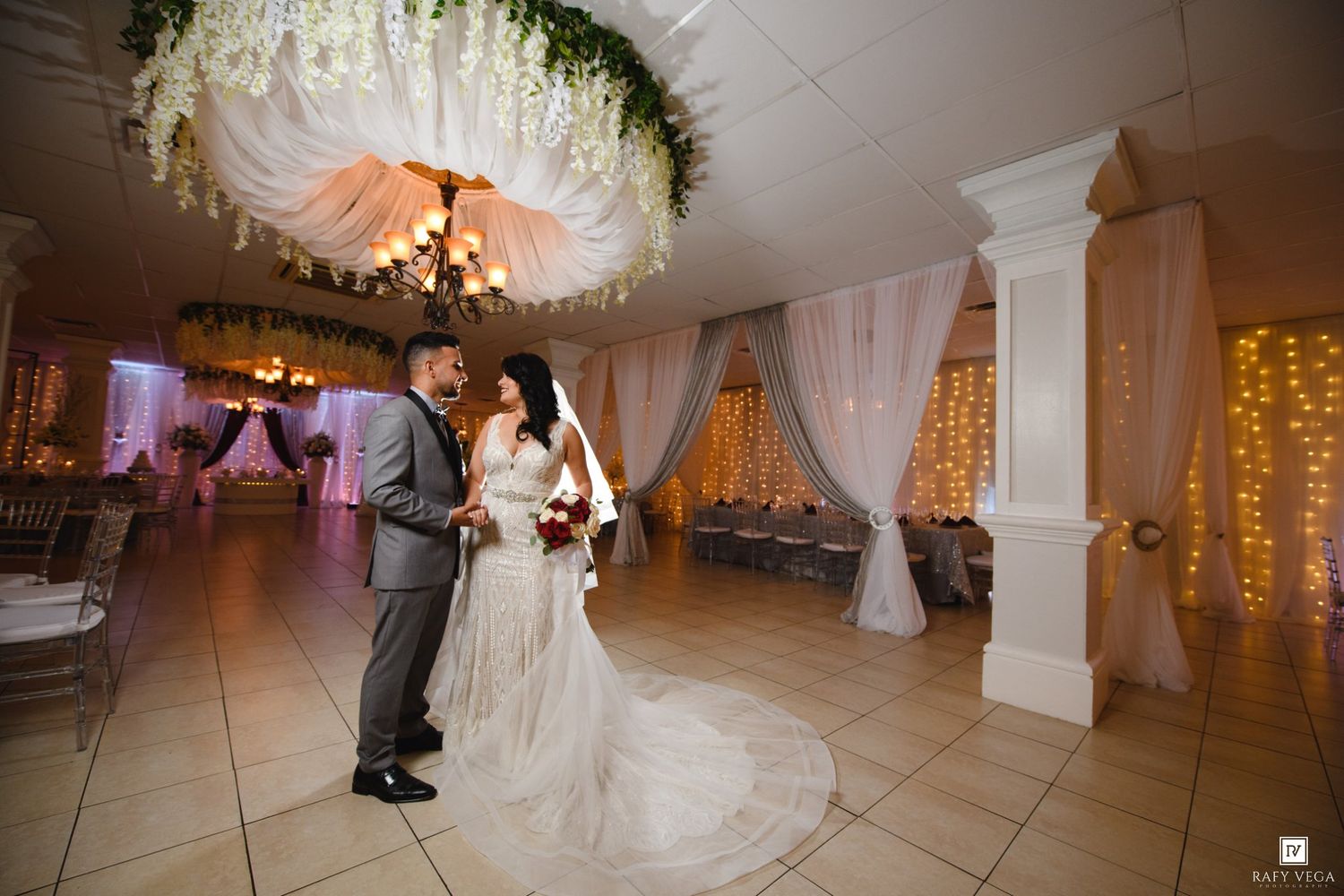 Bride and groom share a romantic first dance in an elegantly decorated wedding venue with draped fabric and twinkling lights.