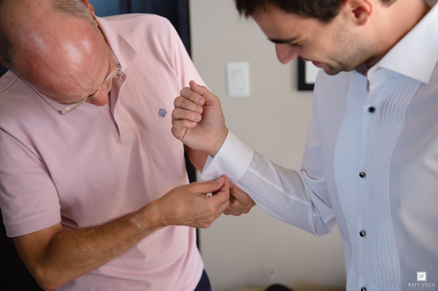 Older man helping young groom fasten cufflinks on white dress shirt before wedding ceremony.